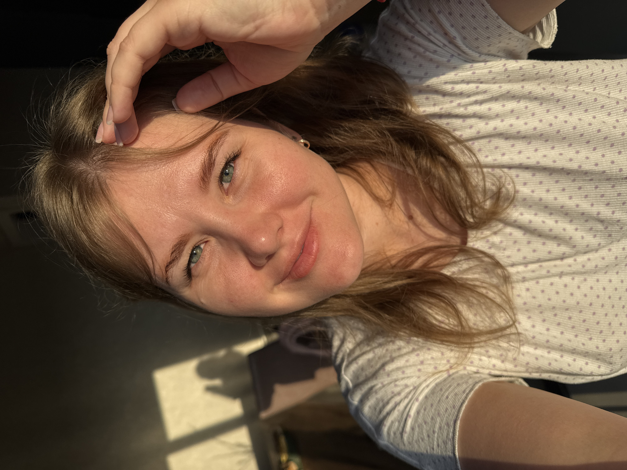 A close-up, slightly angled portrait shows a young woman with fair skin, light brown hair, and green eyes. Her left hand is raised towards her forehead, fingers gently touching her hair. She is wearing a light-colored long-sleeved shirt with small, evenly spaced purple dots. The background shows a shadow cast on what seems to be a flat surface or floor, with a bright rectangular patch of light. The image could have been taken indoors near a window. The location, Ферма, Russia, is not discernible from the visual information present in the image.