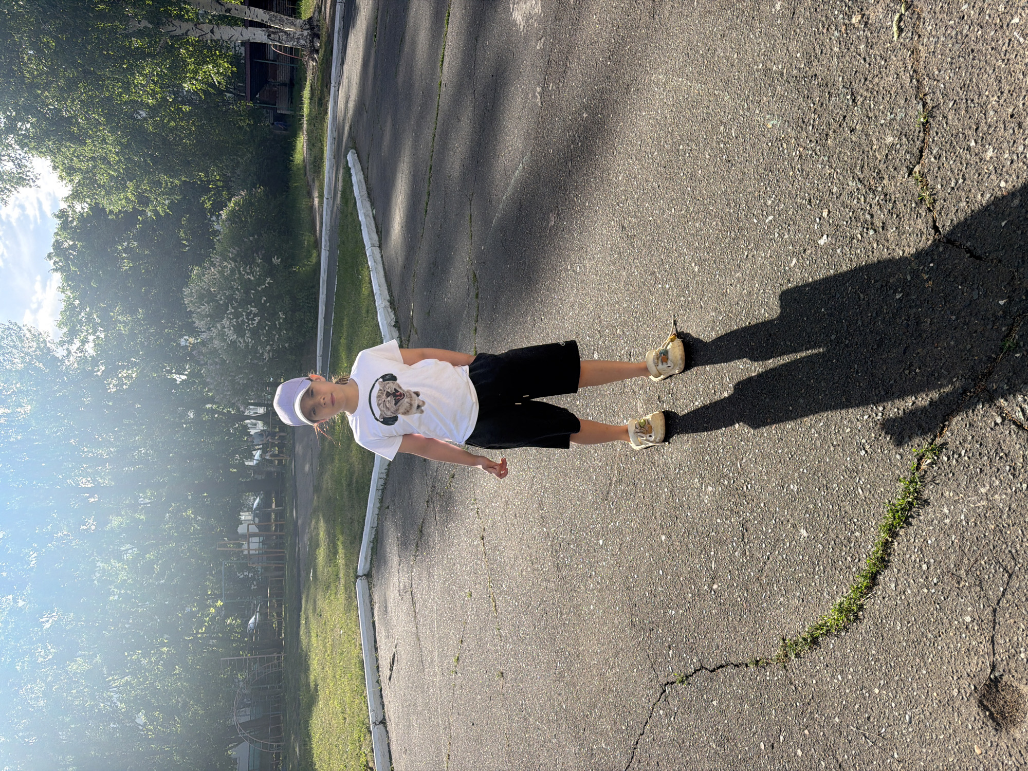 A person stands on an asphalt surface with cracks and patches of grass in Chaikovsky, Russia. The person wears a white t-shirt with a graphic print, black shorts, and light-colored sneakers. A light purple baseball cap is on their head. The sun casts a shadow of the person to the right. To the left of the asphalt surface is a grass area with a concrete border, and trees are visible in the background. A building or structure is partially visible between the trees. The time of day appears to be daytime, likely sunny. A play ground with play equipment is visible behind the trees.