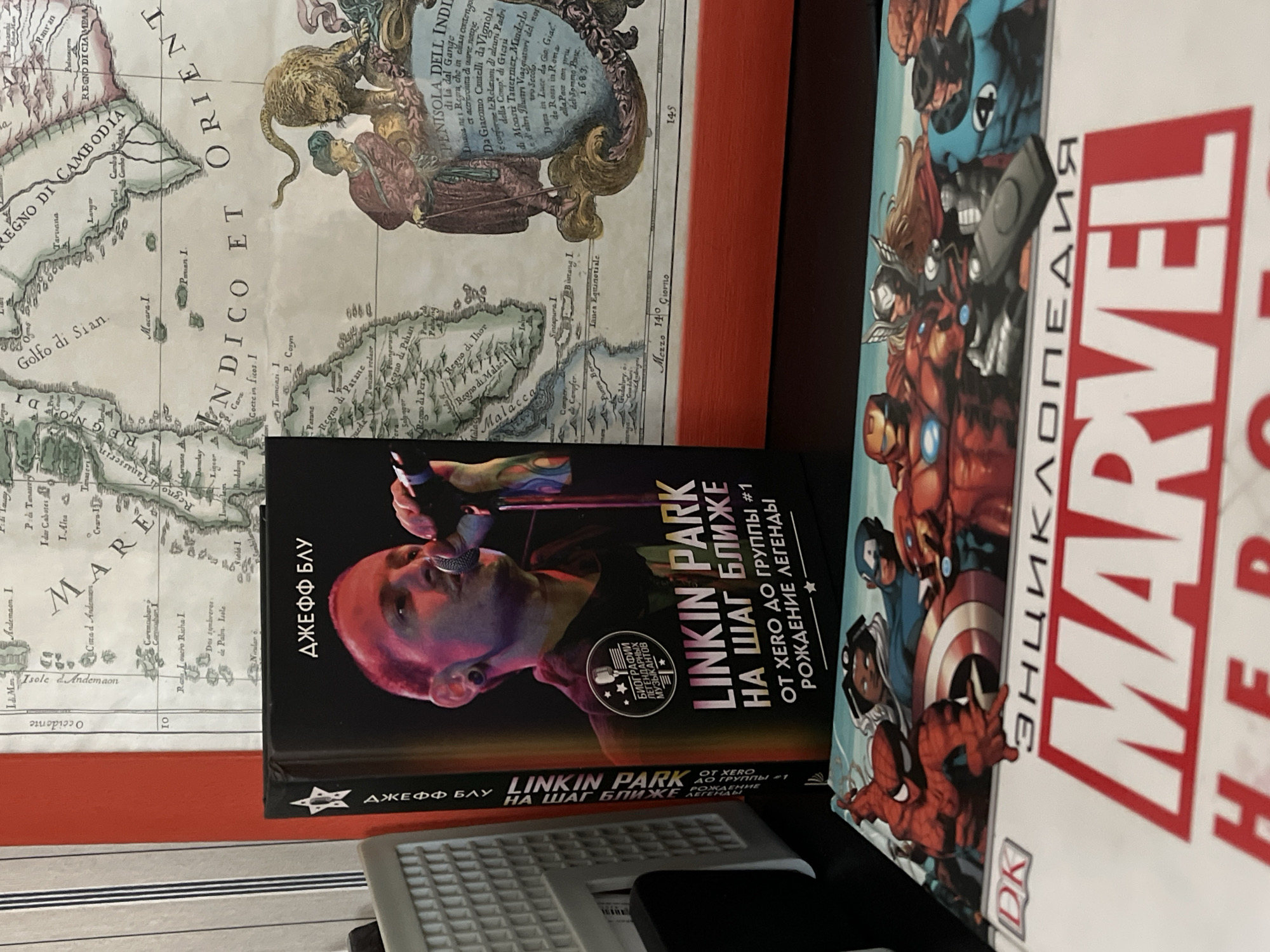 The image shows a close-up of several objects lying on what appears to be a desk or table. From left to right, there's a framed map, a black book titled "LINKIN PARK" in Russian, and a "MARVEL" encyclopedia. The map is old and shows a portion of Southeast Asia. The Linkin Park book features a picture of Chester Bennington on the cover and is written in Russian. The Marvel encyclopedia features a red and white title on its cover, also in Russian, with comic book characters visible. The background shows horizontal blinds. The image does not reveal any identifiable information to infer if it was taken in the city of Krasnoyarsk, Russia.