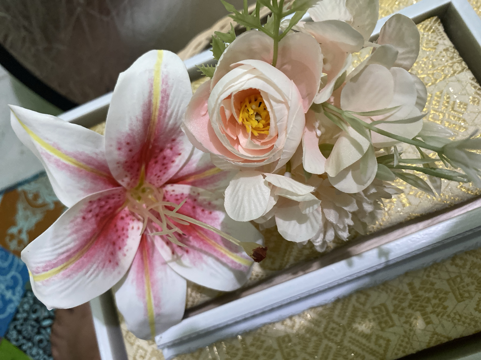 A close-up, overhead shot captures an arrangement of artificial flowers within a white box. The prominent flower in the upper portion of the frame is a large, open lily with white petals streaked with vibrant pink and delicate yellow accents. Its stamen are visible and pale yellow. In the lower section, a cluster of smaller, soft pink roses and white flowers with green leaves creates a softer, more delicate visual. The flowers rest on a textured, golden fabric with an intricate pattern, likely silk or brocade, which fills the base of the box. The background is blurred but suggests a colorful, patterned surface and possibly a brown, textured mat. The image evokes a sense of decoration or gift-giving, typical of events or displays. No people or specific locations within Kuala Pembuang, Indonesia, can be identified from this image.