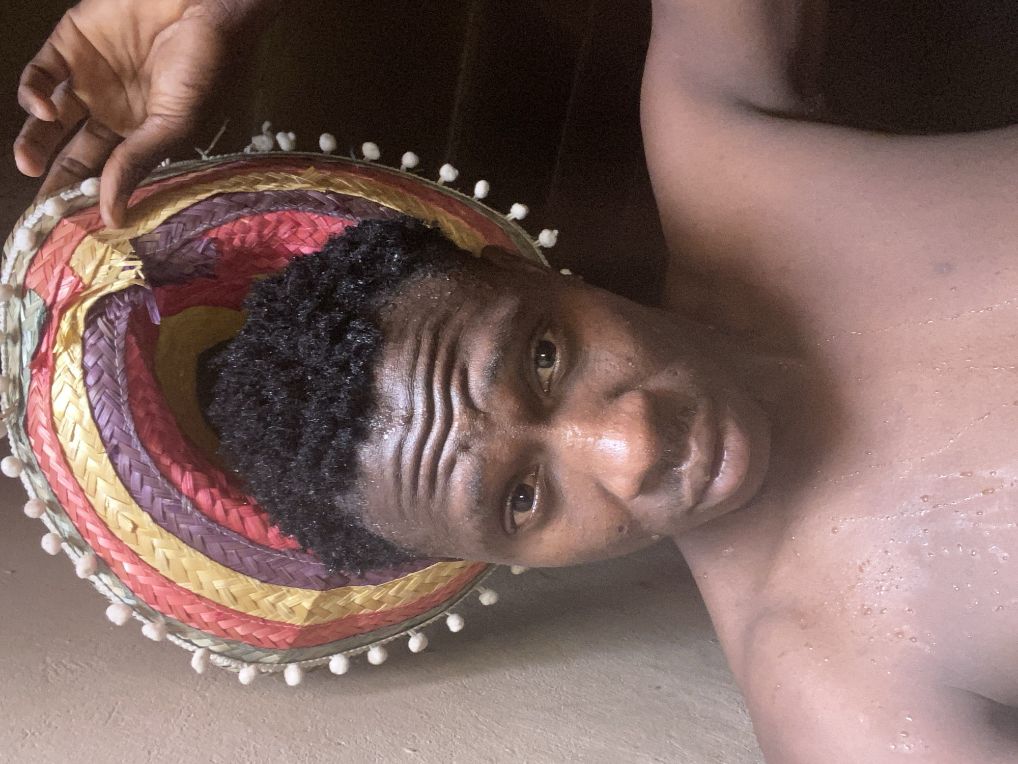 A close-up, angled shot of a dark-skinned man. His face appears sweaty. A colorful, straw-like hat with white pom-poms around the edge rests on his head and is held by his hand. The background is dark and indistinct. There is no indication of Bukuru, Nigeria.