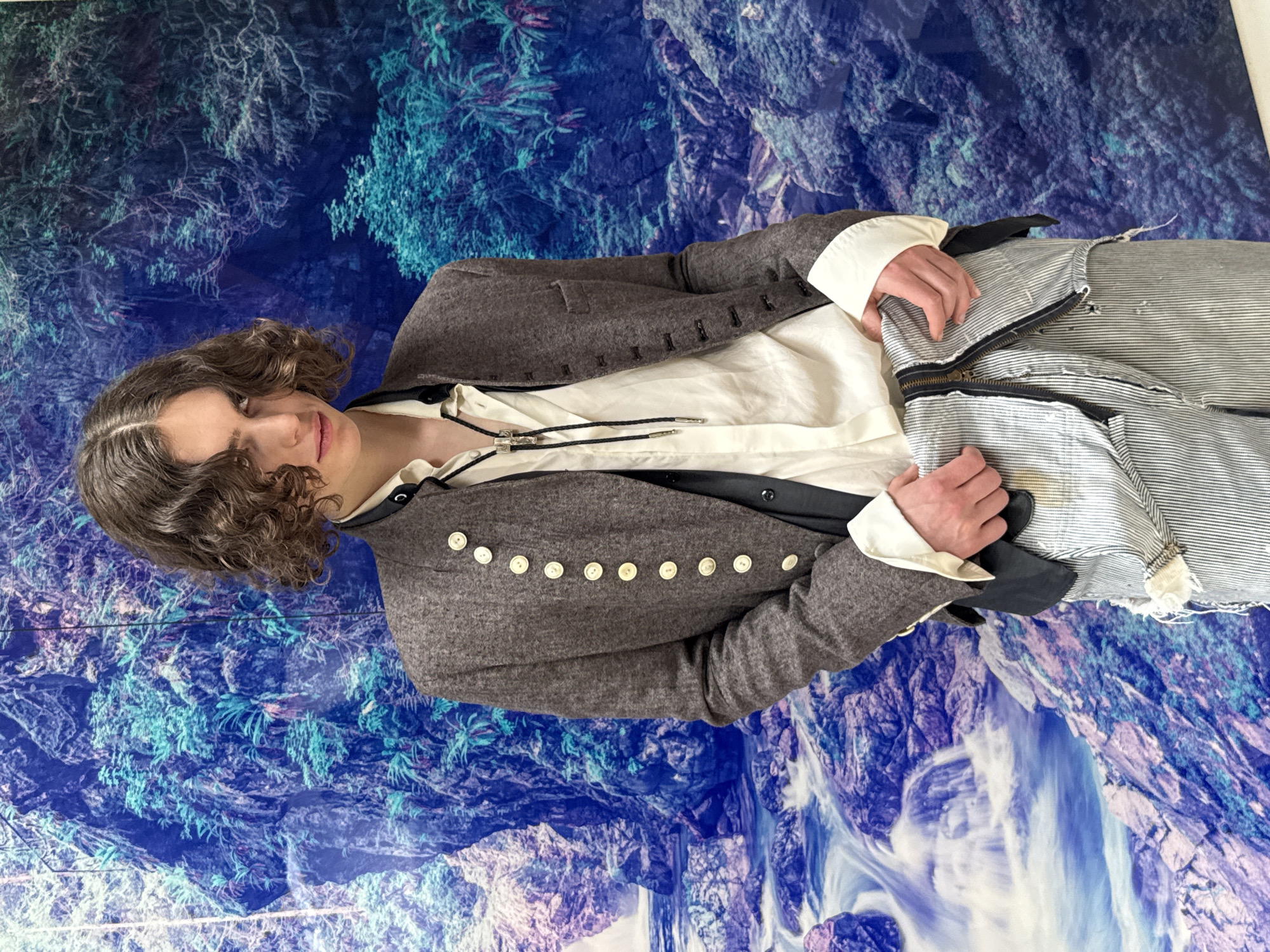 A person with curly, dark hair is wearing a gray jacket with white buttons, a white shirt with a black bolo tie, and striped pants with a visible zipper. They are standing or posing against a background that appears to be a mural or a large-format print depicting a landscape with blue and white elements. The background is abstract. The person is holding the striped pants in their hands.
