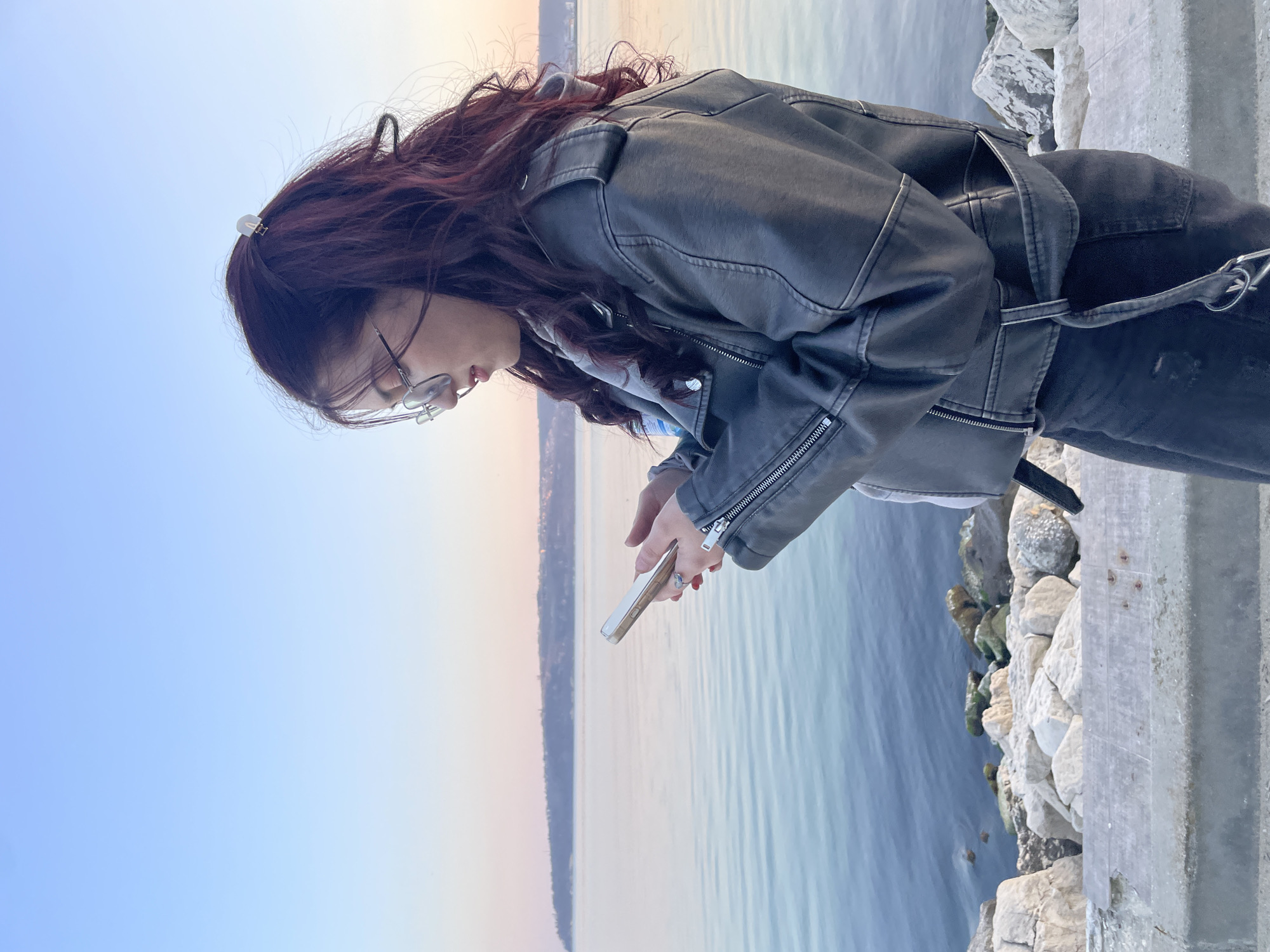 A woman with dark red hair stands on a concrete pier or walkway, looking down at a smartphone held in her hands. She wears glasses, a black leather jacket, and dark pants. The background shows a calm body of water, possibly the Black Sea, with a hazy horizon. Based on the water color and visible cityscape, the location is likely in the city of Varna, Bulgaria, situated on the coast of the Black Sea. Rocks are visible below the concrete pier.