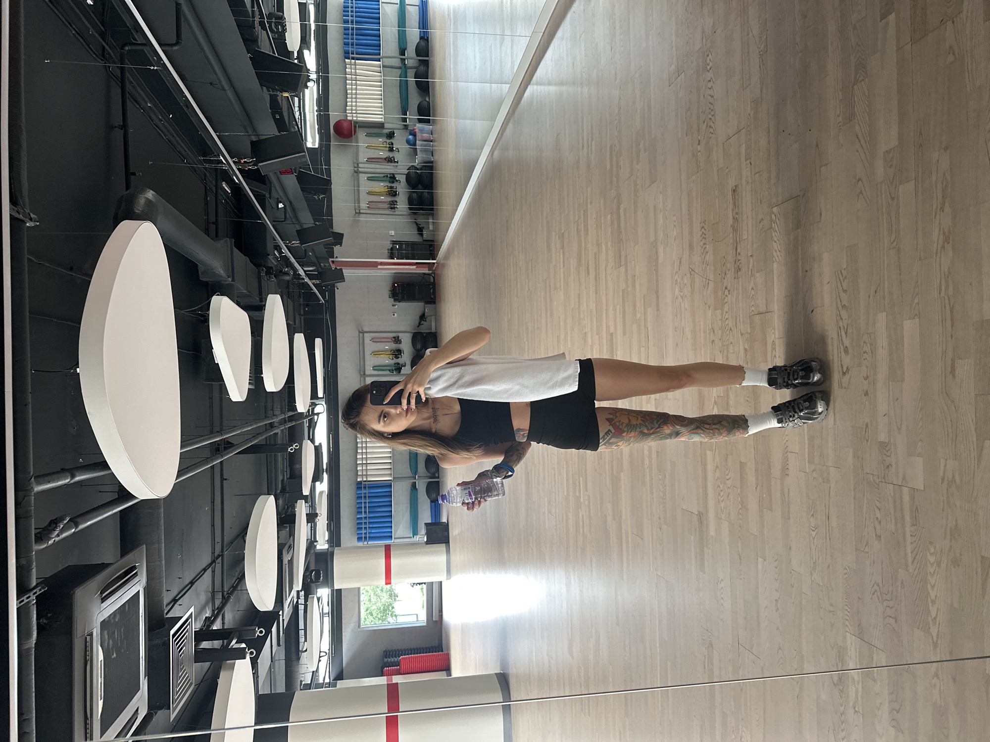A young woman with extensive leg tattoos stands in a gym and takes a mirror selfie. She is wearing a black sports bra, black shorts, white socks, and black sneakers. She holds a clear plastic water bottle in her left hand and a white towel over her right shoulder. The gym has a polished wooden floor, large mirrors on one wall, and athletic equipment visible in the background, including yoga mats and stability balls. The ceiling features industrial elements like exposed pipes and ventilation systems, along with modern, disc-shaped light fixtures. The image is taken from a full-body perspective, with the woman centered in the frame. There is no information available to identify the city as Moscow, Russia.