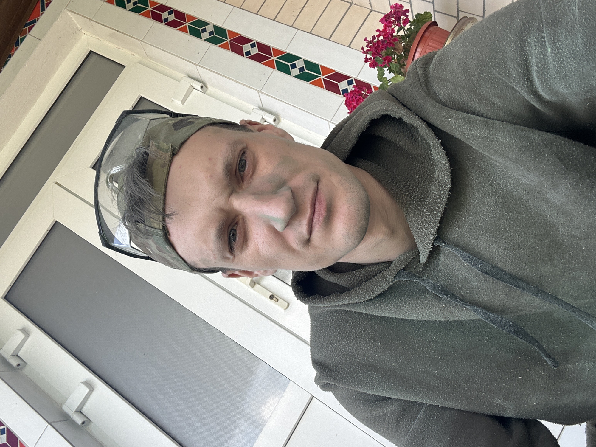 A young Caucasian male, appearing to be in his late 20s or early 30s, is captured in a selfie. He has short, dark brown hair, a strong jawline, and is looking directly at the camera with a neutral expression. He is wearing a dark olive-green hooded sweatshirt with drawstrings and a camouflage baseball cap. Safety glasses are perched on top of his cap. Green face paint is visible on his cheeks and nose, suggesting a possible outdoor activity or preparation for one. The background reveals a building with white trim and a decorative tiled border featuring geometric patterns in red, green, and blue. A potted plant with vibrant pink flowers is situated to the right of the frame, adding a touch of color to the scene. The setting appears to be outdoors, possibly a porch or entrance to a building, with the characteristic architectural style suggesting a location in Ukraine, specifically in the city of Makiv.