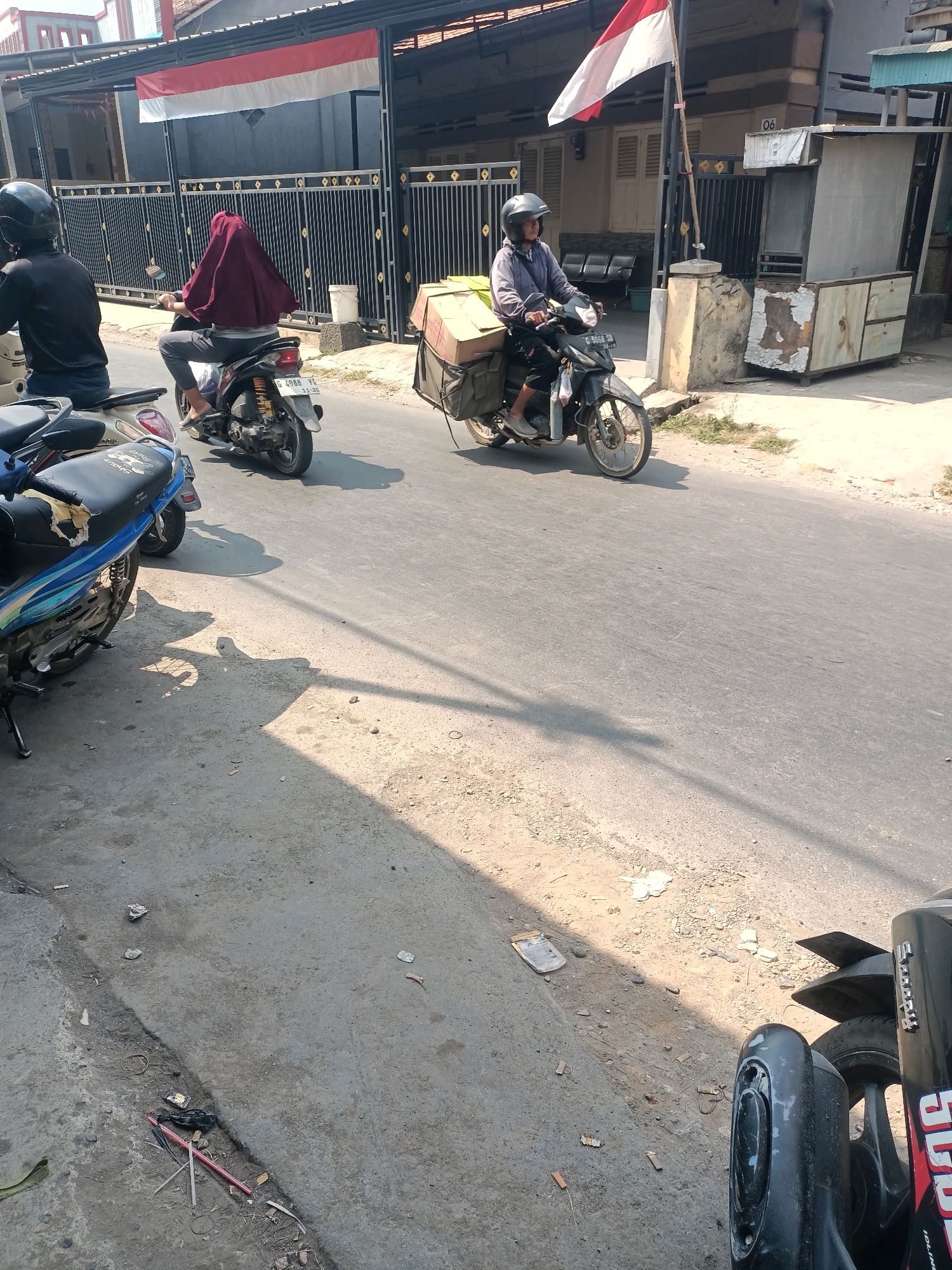 The image captures a street scene in Tembok Banjaran, Indonesia, characterized by a sunny day with distinct shadows cast on the asphalt and roadside. In the foreground, several motorcycles are parked, one of which is a blue and white model with a torn seat. Further down the road, two individuals are riding motorcycles. The first motorcycle, closer to the foreground, is ridden by a person wearing a black helmet and a dark shirt, with a white motorcycle. The second motorcycle, further along the street, is ridden by a person wearing a gray jacket and a black helmet, transporting several cardboard boxes secured to the back. This motorcycle is black. A red, white, and blue flag, likely the Indonesian flag, is prominently displayed on a pole near a building. The surrounding environment includes a building with a dark fence, a small shack-like structure, and a paved road with a dirt shoulder. Litter is visible on the roadside.