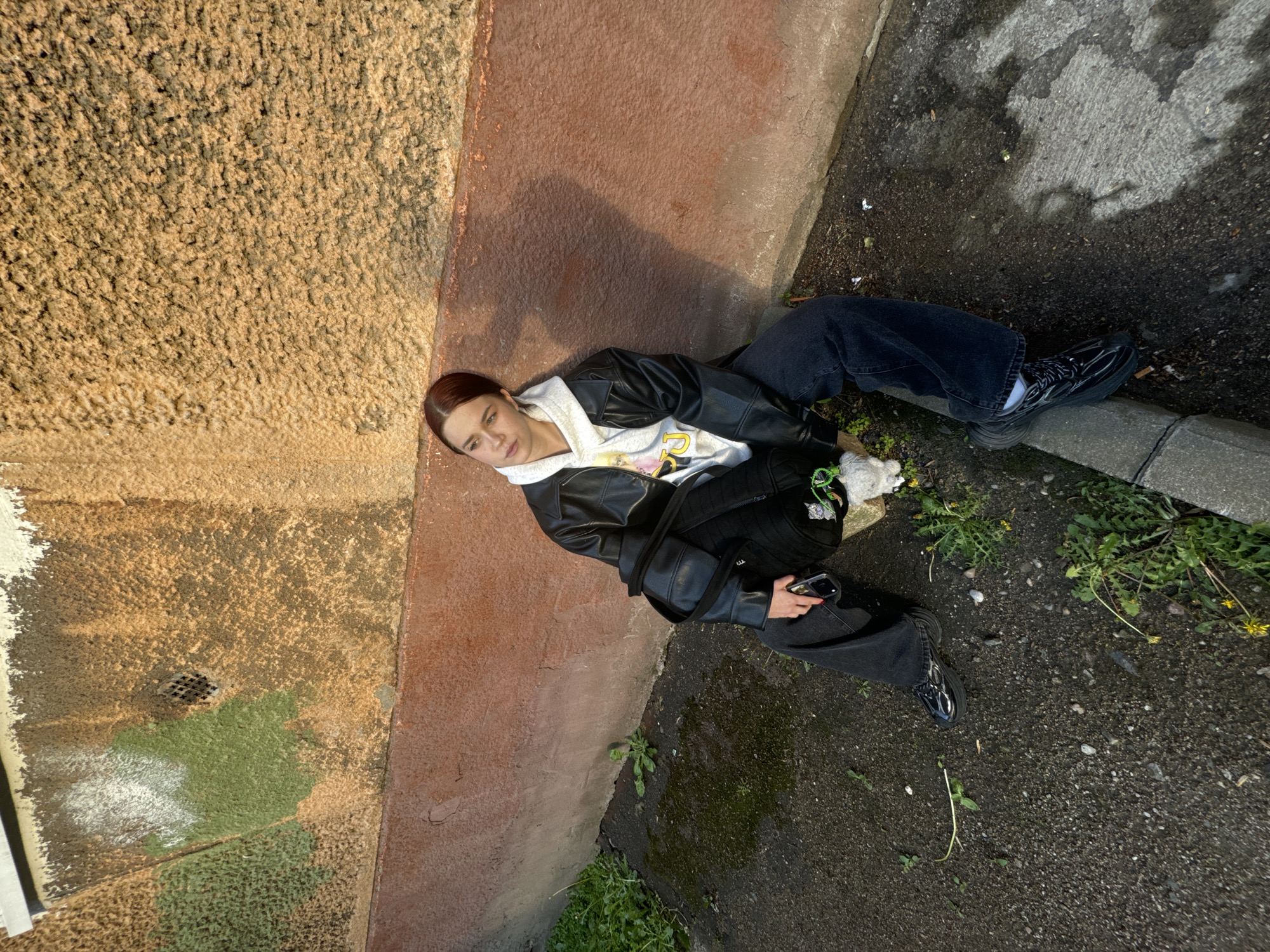 A woman lies on the ground between a wall and a street in Unknown, Unknown. The wall has two textures and colors, one yellow and stucco-like, and the other red. The street is asphalt. The woman wears black pants, a black leather jacket, a white hooded sweatshirt, and black shoes with white socks. She lies between the wall and the street. She holds a phone in one hand. Small green plants grow on the edge of the street.
