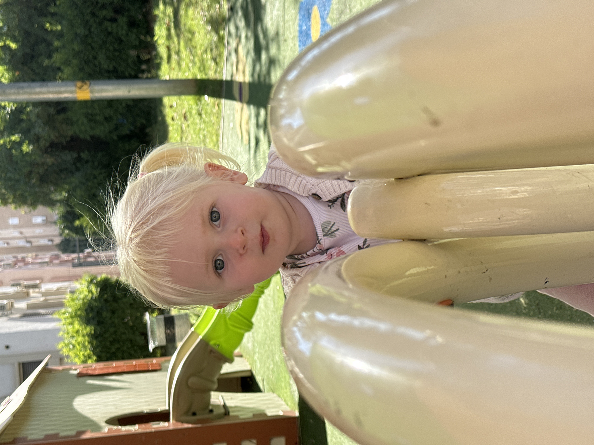 The original image, rotated 90 degrees clockwise, depicts a close-up of a young child in a bright outdoor setting, likely a playground in Petah Tikva, Israel.
The main subject is a young girl with fair skin, light blonde hair tied in a ponytail, and striking blue eyes. She looks directly at the camera with a neutral to slightly curious expression. She is wearing a light-colored long-sleeved top with a subtle leafy or floral pattern, layered under a light pink or beige knitted cardigan.
She appears to be playing on a playground structure, with large, smooth, cream-colored or light yellow curved bars prominently in the foreground, partially framing her face. Behind her, parts of a green slide and a small wooden playhouse are visible.
The background shows a sunny day with green grass and foliage from trees or bushes, indicating an outdoor park environment. Shadows are cast on the ground, confirming bright daylight. Further in the background, out-of-focus residential buildings can be seen, suggesting an urban or suburban park setting. A metallic pole with a yellow marking is also visible among the greenery. No discernible text is visible in the image.