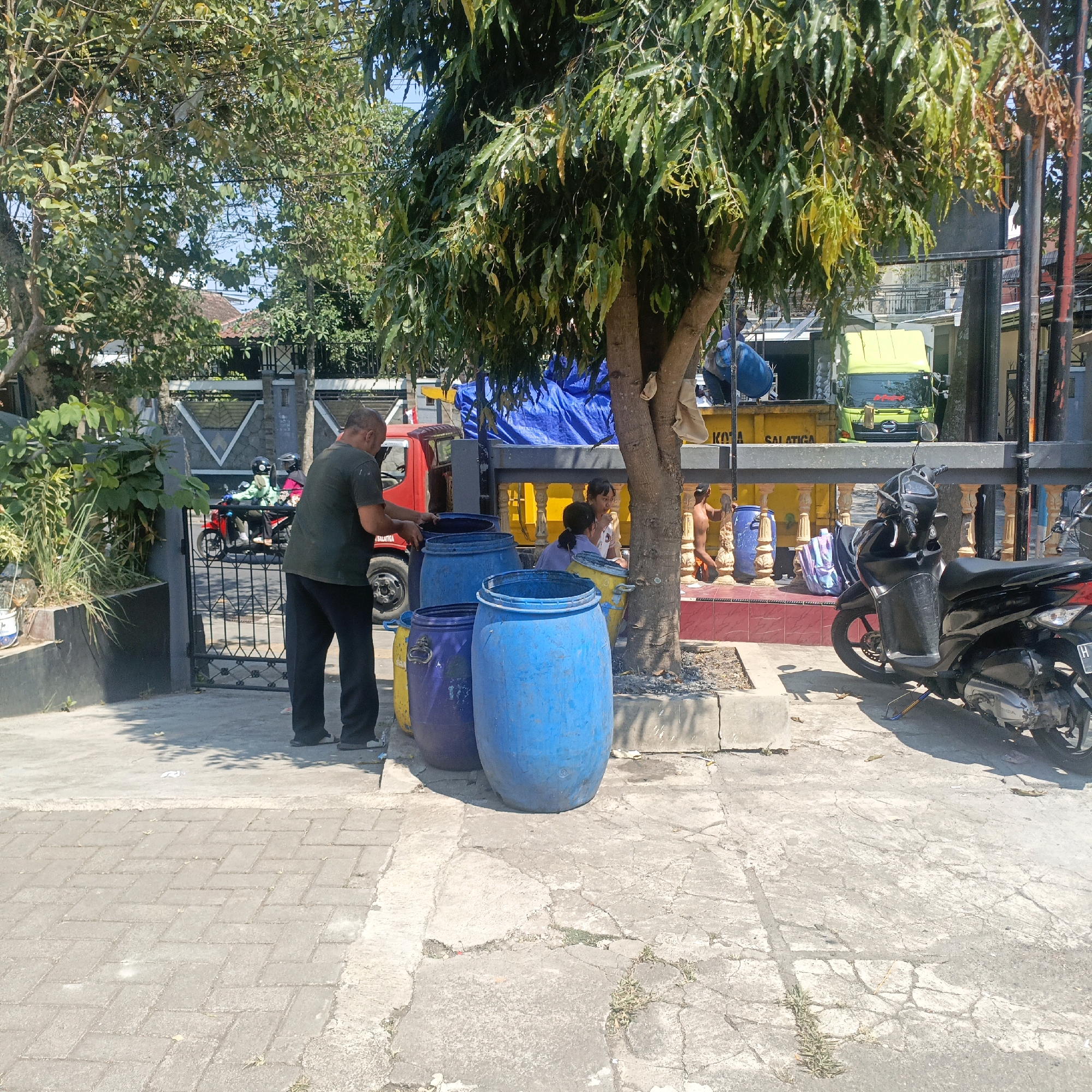 In Salatiga, Indonesia, a man in a dark shirt and pants stands beside a row of large plastic barrels—one purple and two blue—on a paved area. He is facing away from the camera and appears to be interacting with one of the blue barrels. Behind him, a red truck is visible, and beyond that, a yellow truck with a blue tarp covering its load is parked. Several people are seated or standing near the yellow truck. In the foreground, a black motorcycle is parked on a cracked concrete surface. A metal fence with decorative elements runs along the background, separating the street from the area where the trucks and barrels are situated. Green trees with abundant foliage provide shade over the scene, with the bright sky visible through the branches.