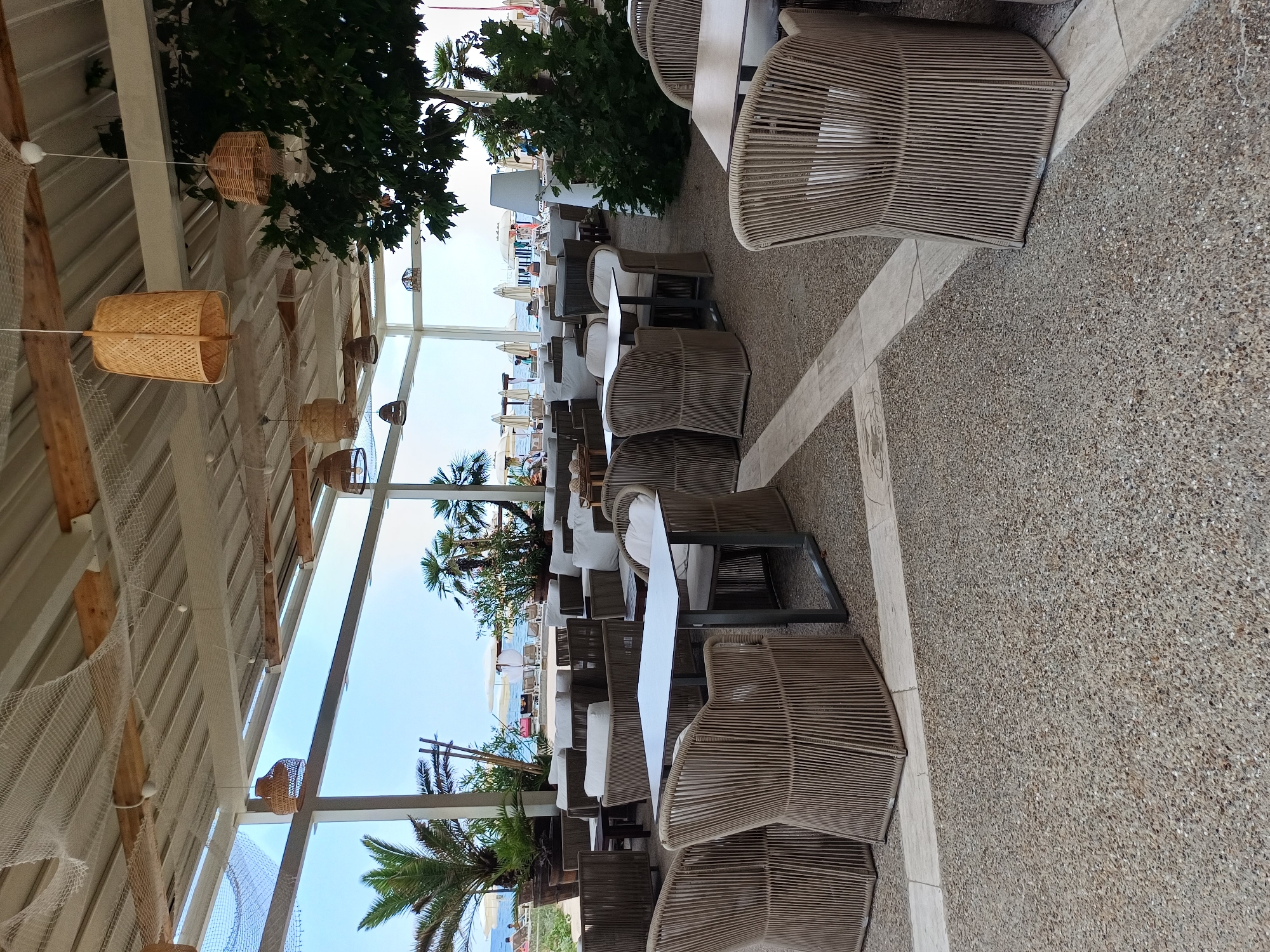 The image depicts an outdoor seating area of a restaurant or beach bar in Varna, Bulgaria, situated near the coast. The scene is an open-air pavilion with a sloped, white-paneled roof supported by white beams. White fishing nets are draped across the ceiling structure, adding a decorative, nautical theme. Hanging from the ceiling are several woven bamboo pendant lamps, some large and conical, others smaller and bell-shaped.
The seating area is furnished with multiple tables and chairs. The tables are mostly square with white tops and dark metal frames. The chairs are a prominent feature, constructed from a woven, light brown material that appears to be rattan or a similar synthetic fiber. They are designed with a rounded, almost basket-like form and have light-colored cushions. In the foreground, a square table is surrounded by four such chairs. Further into the pavilion, more tables and seating arrangements, including some sofa-like structures with cushions, are visible.
The floor is a textured surface composed of small, gray pebbles with some larger, light-colored rectangular tiles interspersed, creating a pathway or pattern.
Beyond the covered seating area, the scene opens to a view of the sea. Palm trees and other lush green vegetation are visible, some in large potted planters. In the distance, beach umbrellas and people can be seen on the sand and in the shallow water, indicating a beachside location. The sky is a clear, pale blue. The overall atmosphere suggests a relaxed, seaside dining or lounging environment. The specific location within Varna is a coastal establishment, likely part of a resort or beachfront complex.