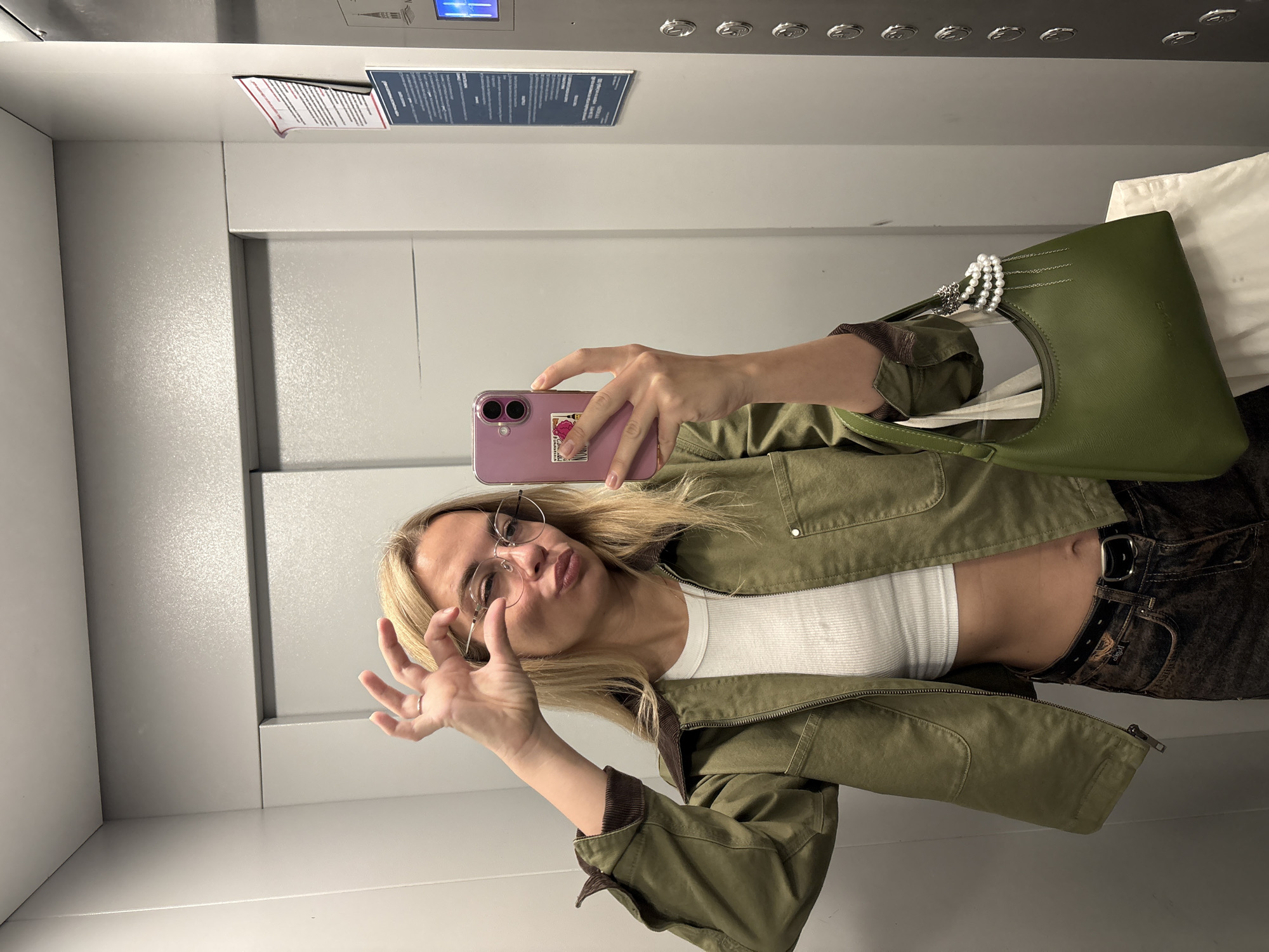 A young woman is taking a mirror selfie in an elevator, appearing to be in Krasnoyarsk, Russia. She is wearing a green jacket, a white crop top, and brown jeans. She has blonde hair and is wearing glasses. Her left hand is raised, and she is making a "peace" sign with her fingers near her eye. She is holding a pink cell phone in her right hand. A green handbag with pearl and chain embellishments hangs from her left arm. The elevator walls are a light gray color, and there is a notice posted on the right-hand wall.