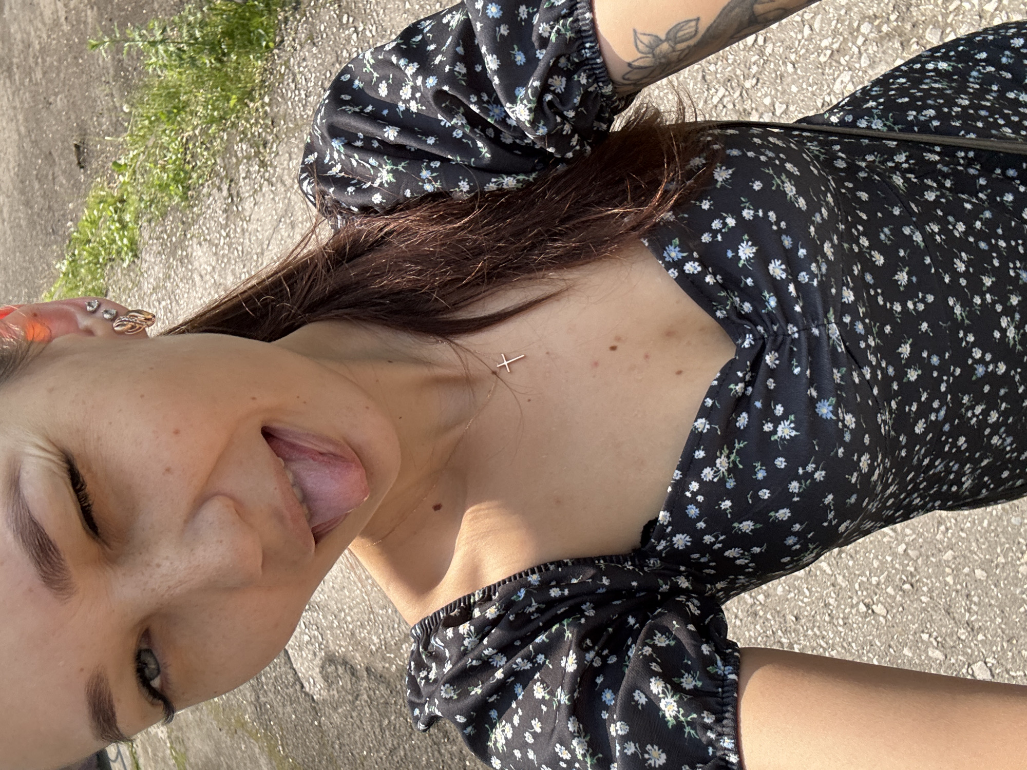 The image shows a young woman with brown hair pulled into a ponytail, wearing a black floral-print dress with puff sleeves and a sweetheart neckline. She is winking and sticking her tongue out at the camera. She has several piercings in her left ear and a cross necklace. A tattoo of a flower is visible on her right arm. The background appears to be an outdoor setting with an asphalt surface and some sparse green vegetation. Based on the style of clothing and the outdoor environment, this appears to be a casual, daytime photograph. Without further information, it is impossible to determine the specific location within Novokuznetsk, Russia, or any specific details about the woman's actions or interactions beyond what is visually evident.