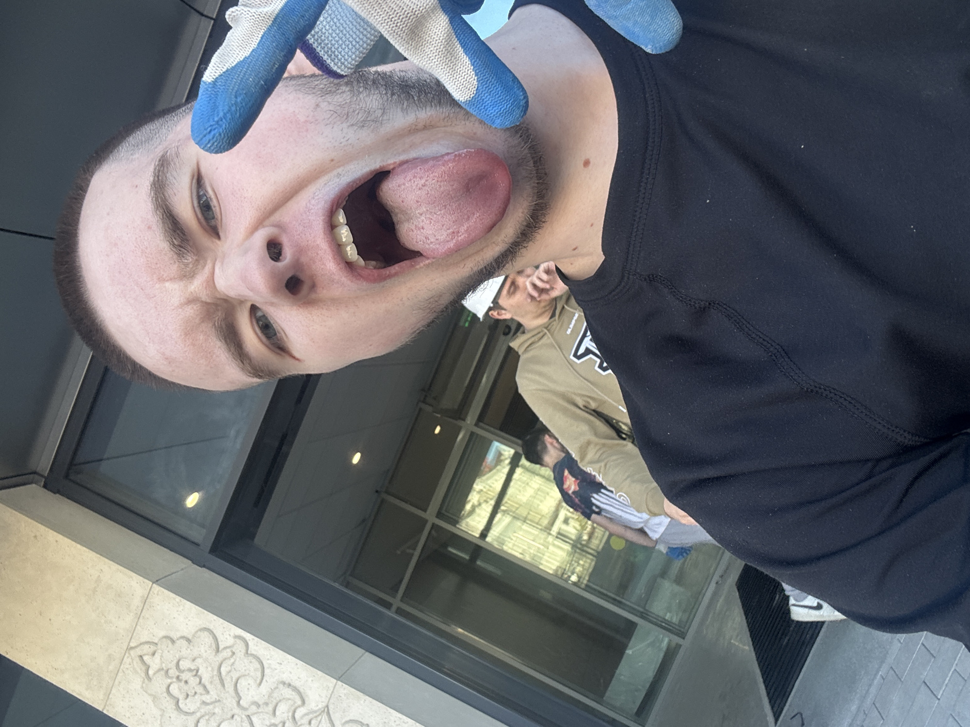 A close-up, tilted selfie features a young man with short brown hair, light skin, and blue eyes. He is sticking his tongue out with his mouth open wide, and making a silly face with his eyebrows raised. A hand wearing a work glove with blue fingertips is visible near his face. He's wearing a black long-sleeved shirt.
In the background, partially visible through a glass doorway, are at least two other individuals. One is wearing a khaki-colored hooded sweatshirt with white lettering, and another is wearing a white shirt with striped sleeves.
The doorway is part of a building with stone or concrete elements. Based on the architectural style and design elements visible, the location may be the city of Kazan, Russia.