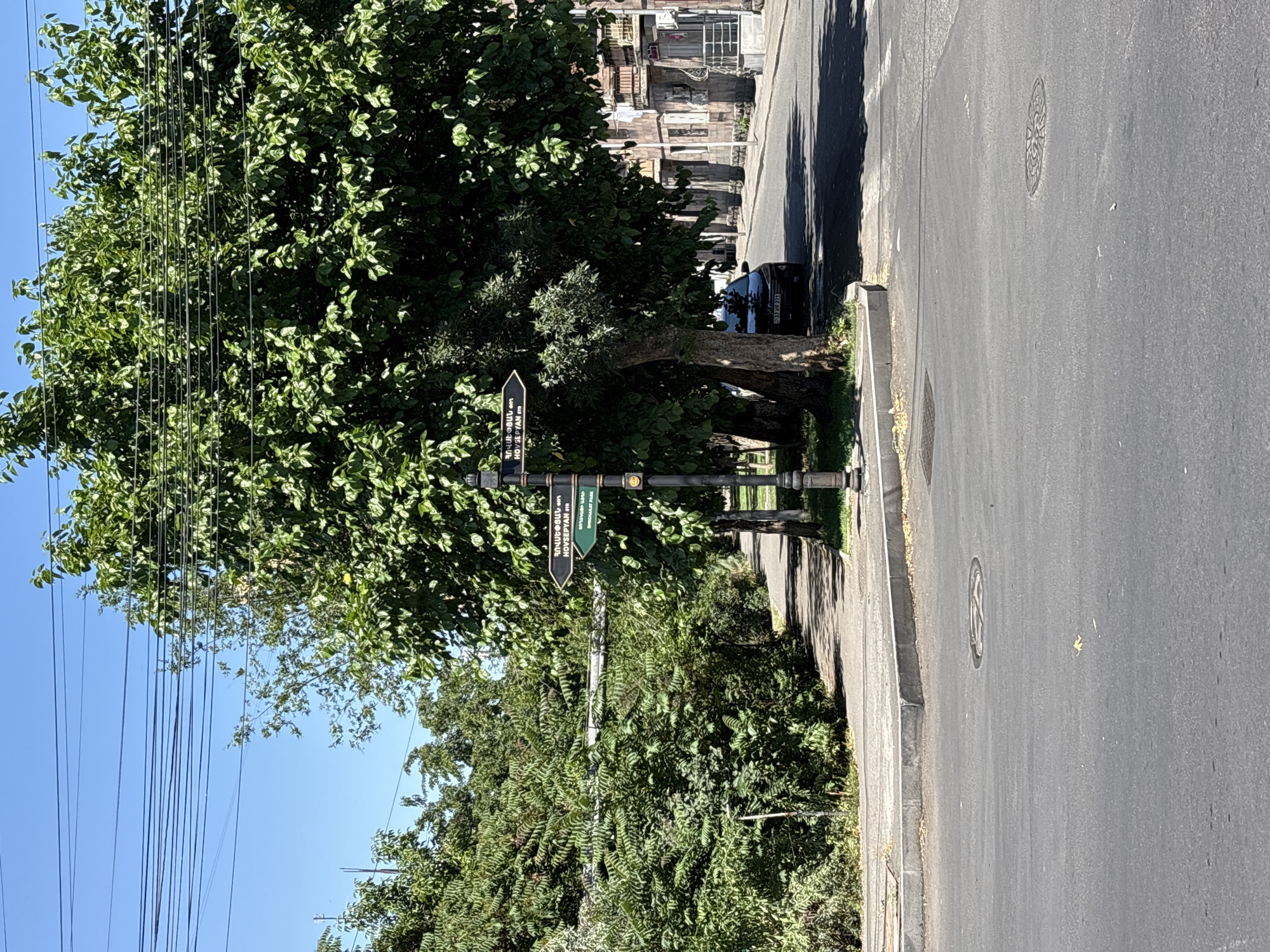 The image depicts a street scene in Yerevan, Armenia. On the left, there is a sidewalk lined with lush green trees and dense foliage. A black street signpost stands prominently, displaying directional arrows with text in Armenian and English. One arrow points left and reads "HOVSEPYAN STR". Another arrow points right and reads "SHAKHRIAR PARK". In the middle ground, a dark-colored car is parked on the right side of the street, partially obscured by trees. The street itself is paved and shows signs of wear. The buildings on the right side of the street are made of stone and have balconies. The sky is clear and blue, indicating a sunny day.