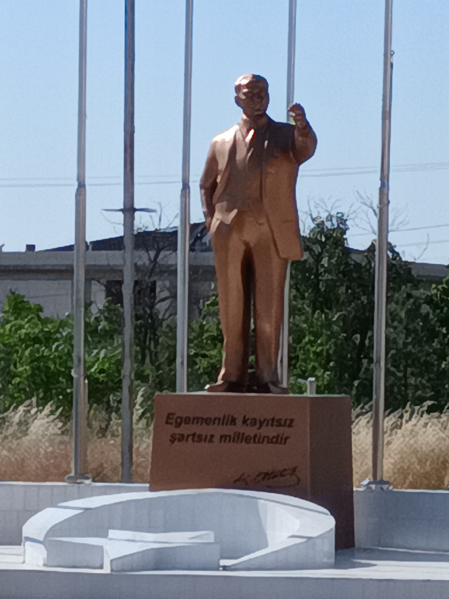 A full-body bronze statue of a man in a suit stands on a pedestal in front of a clear blue sky. The statue faces the viewer, with its right arm raised and its left hand in its pocket. The pedestal is made of red stone and has an inscription in Turkish. Behind the statue are several flagpoles, trees, and a building. In the foreground, there is a white concrete structure with curved edges. The image is taken from a medium distance, capturing the full statue and its immediate surroundings. The overall scene suggests an outdoor public space, possibly a park or memorial, in Bahşılı Belediyesi, Turkey.