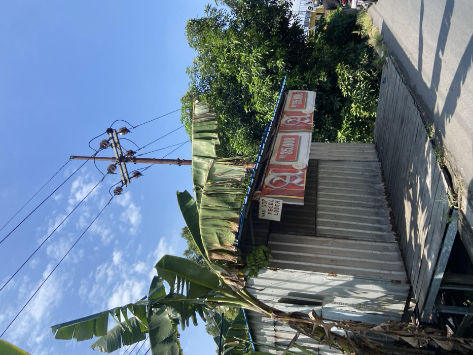 The image shows a small, corrugated metal building with a wooden platform in front of it. The building has a sign above the entrance that reads "Nasi Sop Pecel Lodeh." There are two signs in front of the building with the price "Rp. 35.000" written on them. A banana tree is growing next to the building. A utility pole with several power lines runs through the center of the image. There are some trees behind the building and a road in the foreground. The scene is located at coordinates -7.504 latitude, 112.53 longitude, which is in Mojosari, Indonesia.