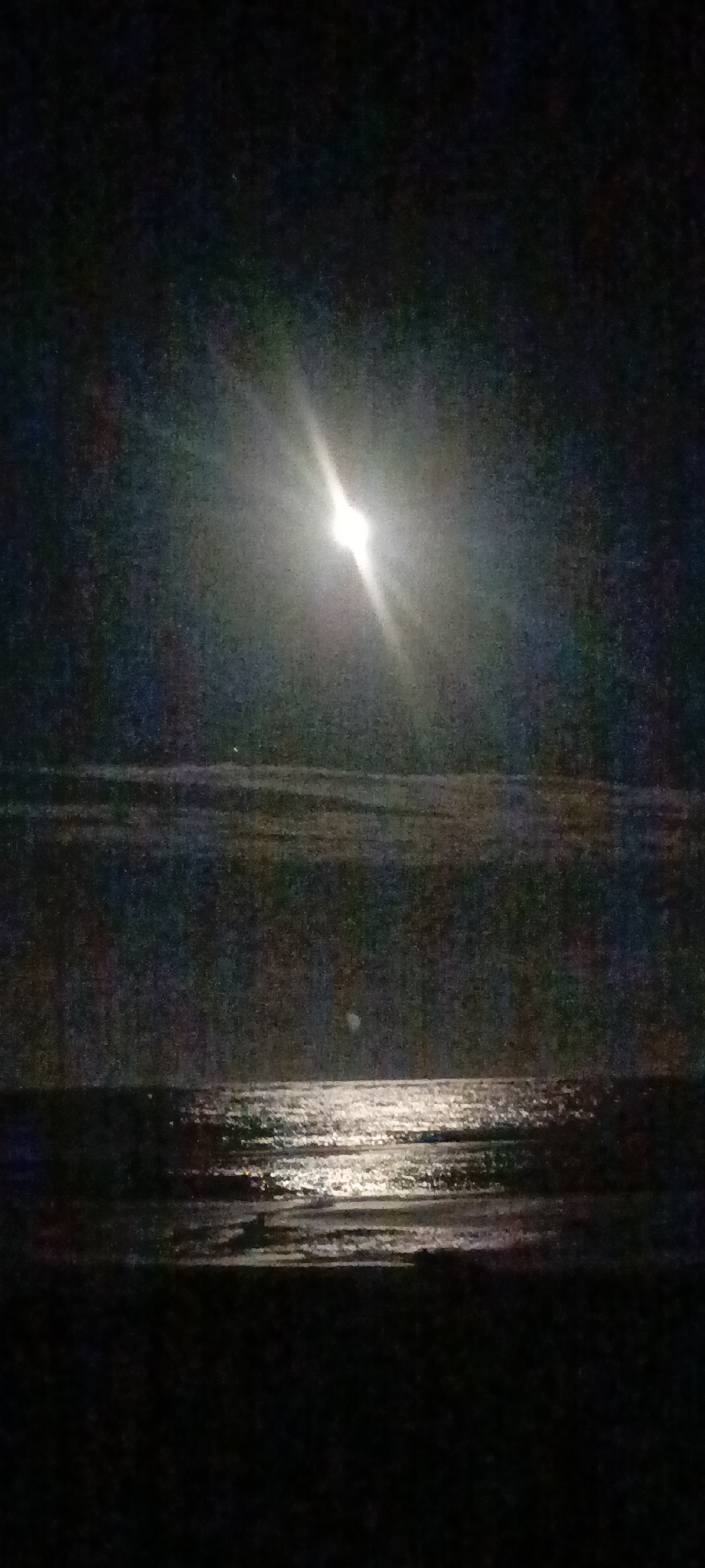 A nighttime photograph captures a moonlit ocean scene. The moon is a bright, slightly elongated disc positioned high in the dark sky, casting a luminous beam that reflects across the water. The water is dark, with distinct, shimmering ripples illuminated by the moonlight. The horizon line separates the water from the sky, and the foreground is a dark, indistinct area, likely the shore. The overall impression is one of tranquility and natural beauty under the night sky.