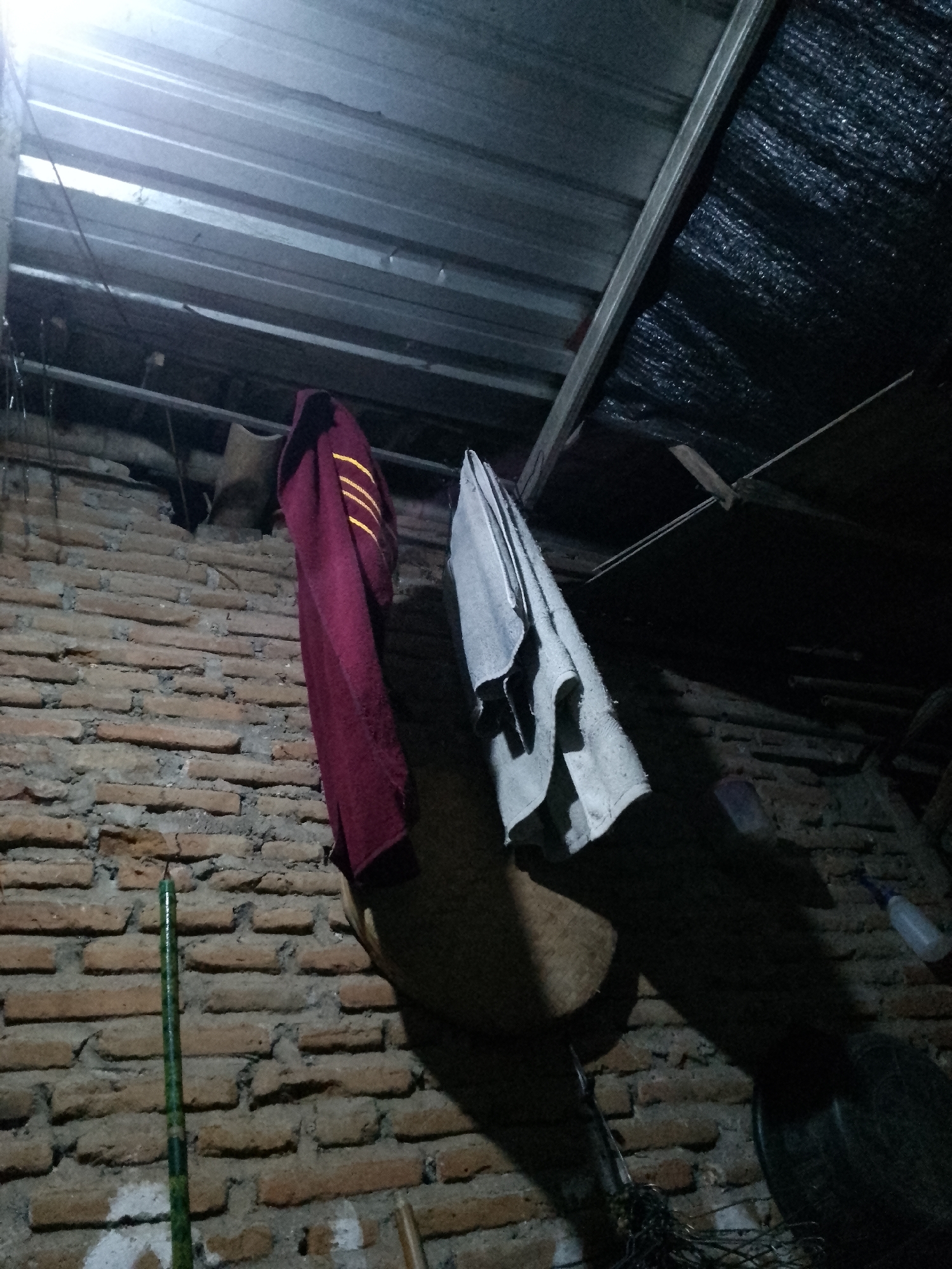 The image depicts an interior scene, likely a storage area or utility space, with a brick wall and a corrugated metal ceiling. Two pieces of fabric are hanging from above. On the left is a dark red garment, possibly a jacket or sweater, with yellow stripes on the sleeves. To its right is a light gray towel. A thin, green, cylindrical object, possibly a stick or rod, is leaning against the brick wall in the lower left quadrant. The environment appears somewhat neglected, with exposed brickwork and what looks like a dark, textured material on the ceiling. The lighting is dim, coming from the upper left, casting long shadows. There are no people visible in the image, and no context is provided about the location within Kauman, Indonesia.
