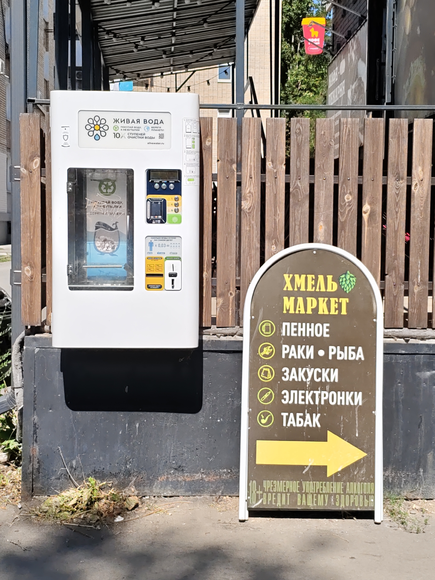 In Rostov-on-Don, Russia, an outdoor scene features a white water vending machine situated next to a wooden fence. The machine has a prominent display with the text "ЖИВАЯ ВОДА" (Living Water) and branding that mentions "10 ступеней очистки воды" (10 stages of water purification). A clear panel on the machine reveals a blue plastic container inside. To the right of the water machine, a brown arched sign with white lettering lists various items: "ПЕННОЕ" (Foamy/Beer), "РАКИ • РЫБА" (Crayfish • Fish), "ЗАКУСКИ" (Snacks), "ЭЛЕКТРОНКИ" (Electronics), and "ТАБАК" (Tobacco), with a large yellow arrow pointing to the right. Below this list, smaller text warns "ЧРЕЗМЕРНОЕ УПОТРЕБЛЕНИЕ АЛКОГОЛЯ ВРЕДИТ ВАШЕМУ ЗДОРОВЬЮ" (Excessive alcohol consumption is harmful to your health). In the background, a building with windows and trees are visible, along with a red and yellow sign for "КОФЕ" (Coffee) or "ПИКАРНЯ" (Bakery) hanging from a structure. The ground in front of the water machine is asphalt with some weeds and debris.