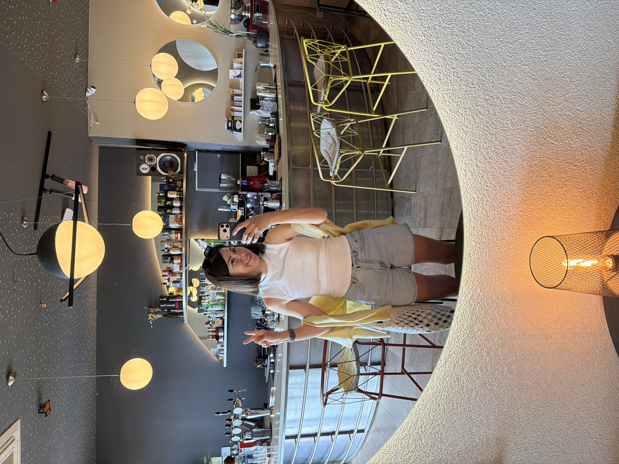 The image shows a woman in a white tank top and grey shorts taking a selfie in what appears to be a bar or restaurant. She is making a peace sign with her left hand. Behind her is a bar with a variety of liquor bottles. There are hanging lights and round mirrors on the wall. The scene includes bar stools and other chairs. The setting is likely Bečići, Montenegro, given the prompt.