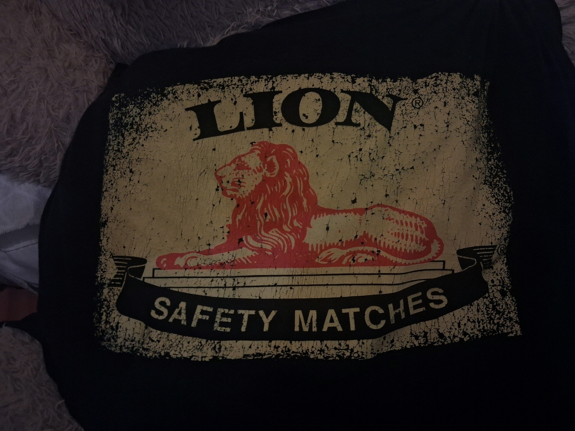 The image shows a close-up of a black t-shirt with a distressed graphic print. The graphic features a stylized illustration of a lion in red, with a mane and body rendered in a somewhat abstract, brushstroke-like style. Above the lion, the word "LION" is printed in large, bold, black, sans-serif capital letters, with a registered trademark symbol (®) to the right of the word. The entire graphic is set within a distressed, textured rectangular frame that appears to be a pale yellow or off-white color, with specks and cracks suggestive of age or wear. Below the lion, a black banner with a distressed texture is draped across the bottom of the frame, displaying the words "SAFETY MATCHES" in white, sans-serif capital letters. The t-shirt is laid out on a textured, furry surface, which appears to be a rug or blanket in a light brown or grey color. The lighting is dim, creating shadows and highlighting the texture of the t-shirt's graphic. There are no people visible in the image. The scene is devoid of any specific landmarks or environmental cues that would suggest a location in Roodepoort, South Africa. The image captures a still, flat lay of the t-shirt.