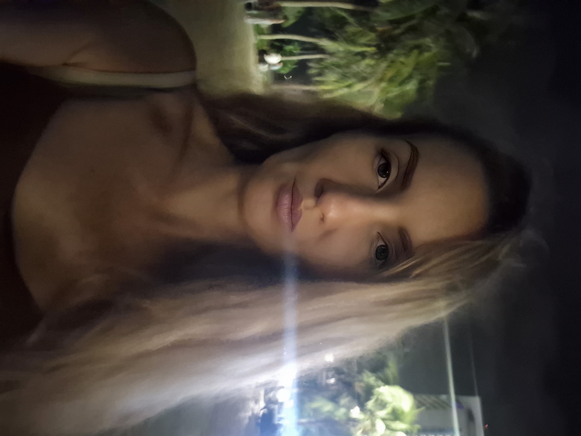 A woman with long blonde hair is taking a selfie outdoors at night. She is wearing a dark top with a light-colored strap visible on her left shoulder. Her facial features are centered in the frame, with her gaze directed towards the camera. In the background, palm trees are illuminated by artificial light, suggesting a tropical or subtropical environment. There are also faint figures of other people in the distance, and a building with lights is visible on the right side of the image. The overall lighting indicates it is nighttime, possibly a beach or resort area. The location is identified as Đà Nẵng, Vietnam.