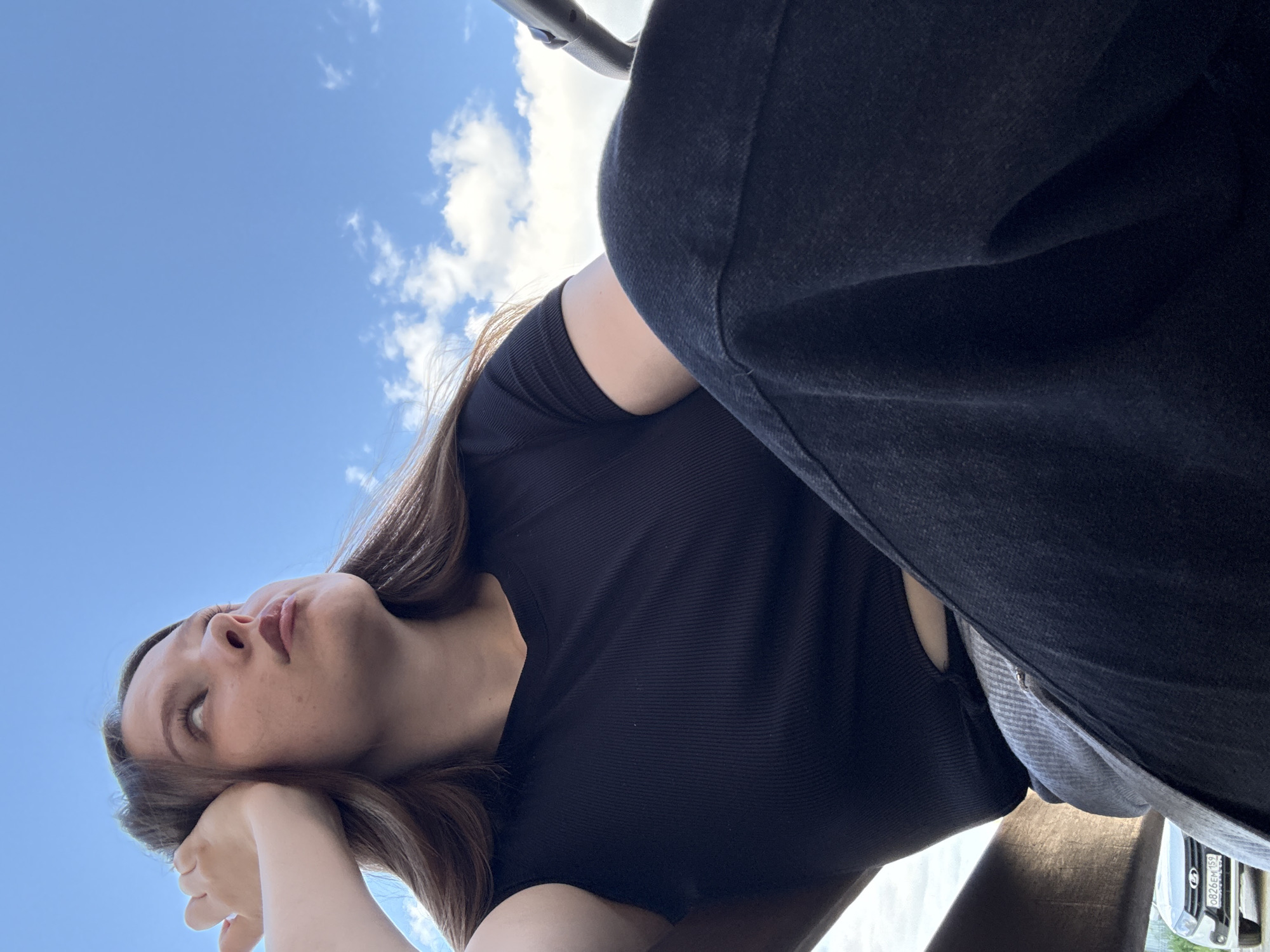 A young individual, appearing female, is depicted from a low-angle perspective, seated outdoors under a clear blue sky with scattered white cumulus clouds. The person has long, light brown hair and is wearing a black ribbed short-sleeved top and dark, likely black, wide-leg trousers. Their left arm is bent, with the hand resting near the left side of their head, and they are looking upwards and slightly to the right of the frame. To the lower left, the front section of a white LADA car is partially visible, displaying the Russian license plate "0826EM 159". The regional code "159" on the license plate indicates registration in Perm Krai, Russia, which encompasses the city of Chaikovsky. A light-colored fence or wall is present in the background behind the car. A dark, indeterminate structure occupies the right side of the frame, possibly part of a vehicle or furniture. The scene is illuminated by daylight.