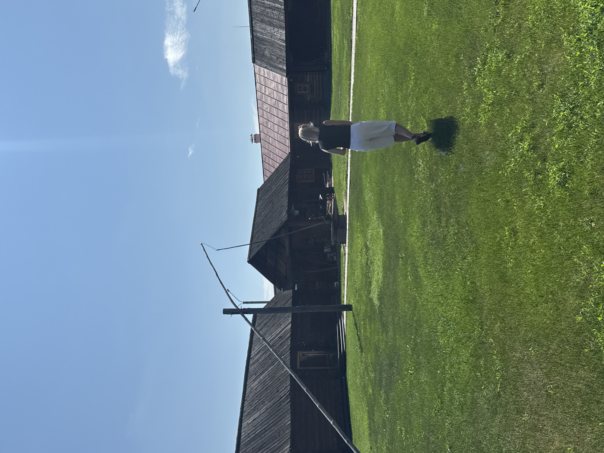 A woman with blonde hair, wearing a black sleeveless top and white shorts, walks away from the camera across a green grassy field. Behind her, a row of dark wooden, rustic buildings with sloped roofs stretches across the background. One building has a red-tiled roof. In the foreground, a wooden well with a long beam is visible, angled towards the left side of the frame. The sky is a clear, bright blue with a few wispy white clouds. The scene appears to be set in an outdoor historical or museum setting, possibly depicting a village in Shushenskoye, Russia.