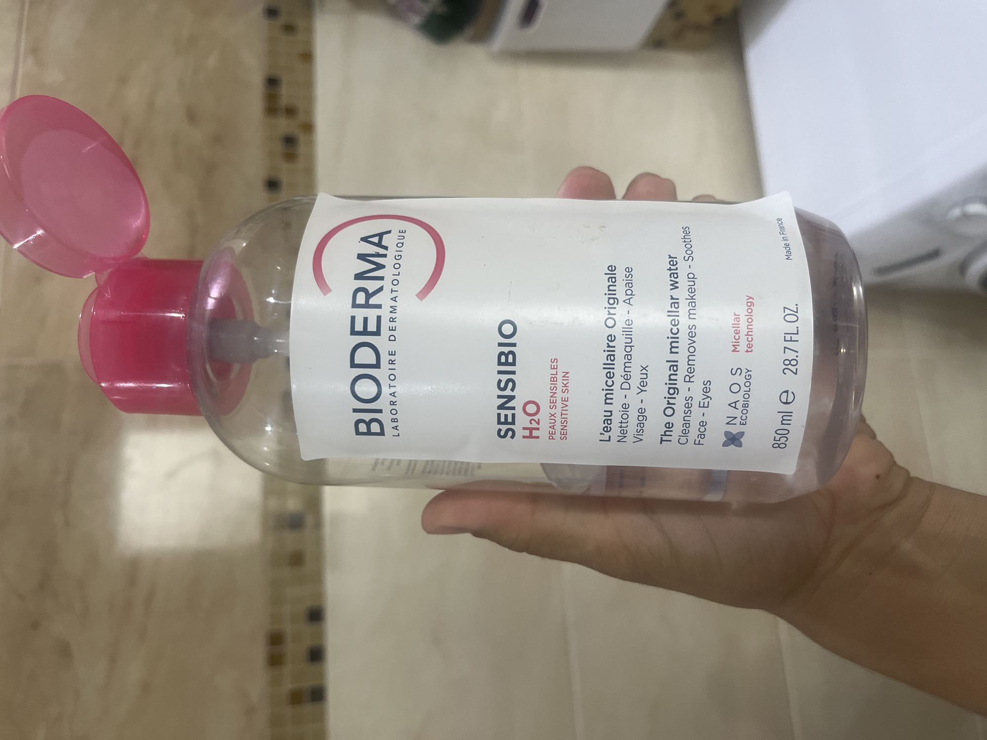A clear plastic bottle of Bioderma Sensibio H2O micellar water is held in a person's hand. The bottle has a pink pump dispenser and a white label with red and black text. The label indicates that it is for sensitive skin, cleanses, removes makeup, and soothes the face and eyes. The bottle contains 850 ml or 28.7 fl oz of product and is made in France. The background appears to be a tiled wall with a mottled beige and brown pattern. The image was taken in a bathroom. There is no indication of the specific location within Onokivtsi, Ukraine.