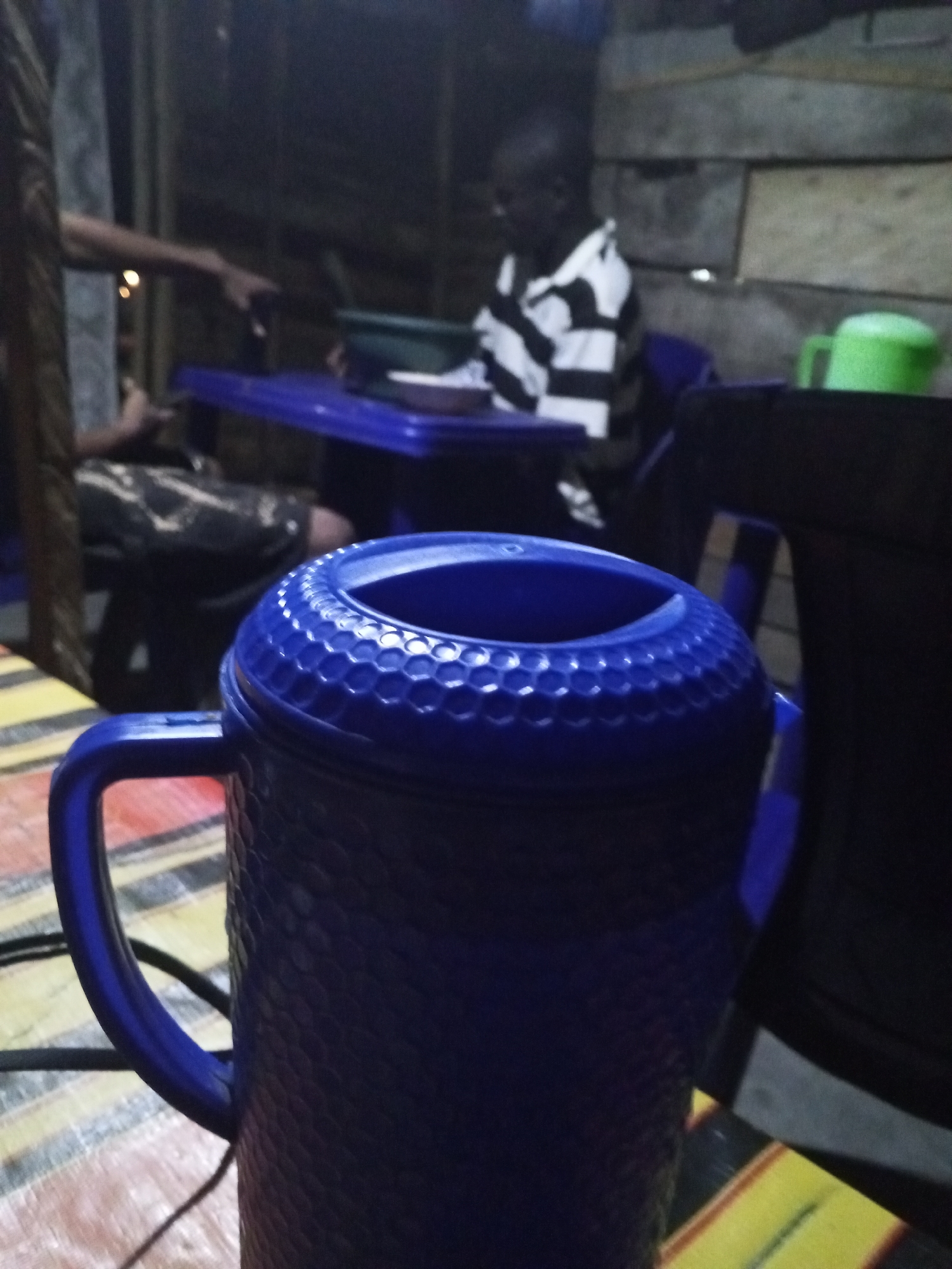 In Lokoja, Nigeria, a close-up shot captures a textured blue thermos with a handle in the foreground. Behind it, a person in a black and white striped shirt sits at a blue table, reaching towards a green bowl. The scene appears to be indoors, possibly at a food stall or restaurant, with wooden walls in the background.