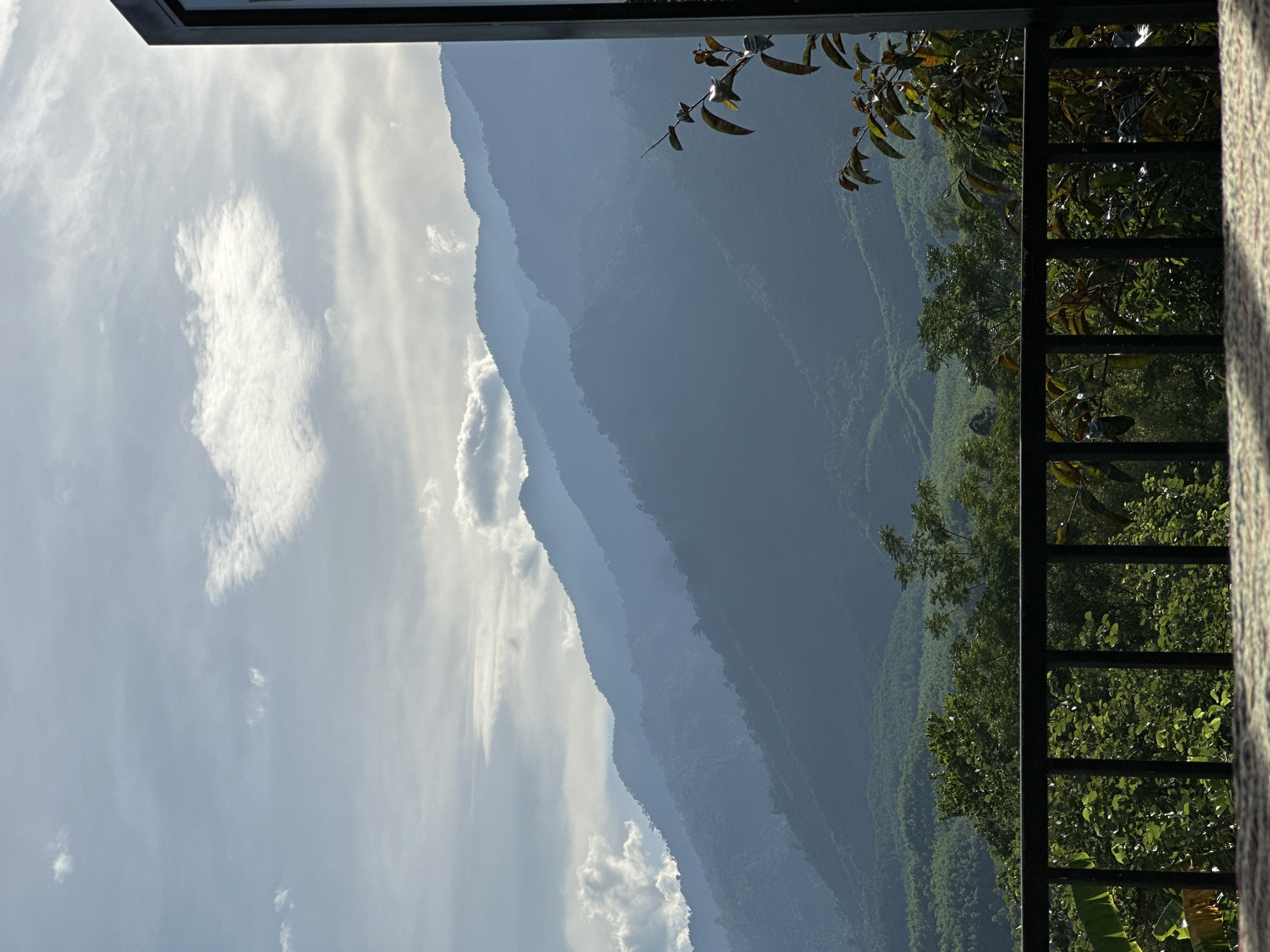 The image depicts a serene mountainous landscape viewed from a balcony, likely in the outskirts of Da Nang, Vietnam. The foreground is dominated by a dark railing, behind which lush green foliage and trees are visible. The mid-ground features rolling hills covered in dense vegetation. In the background, a series of majestic, layered mountains recede into the distance, their blueish hues softened by atmospheric haze. The sky above is a mix of pale blue and white clouds, with sunlight filtering through, creating a gentle, diffused light. The scene evokes a sense of tranquility and natural beauty.