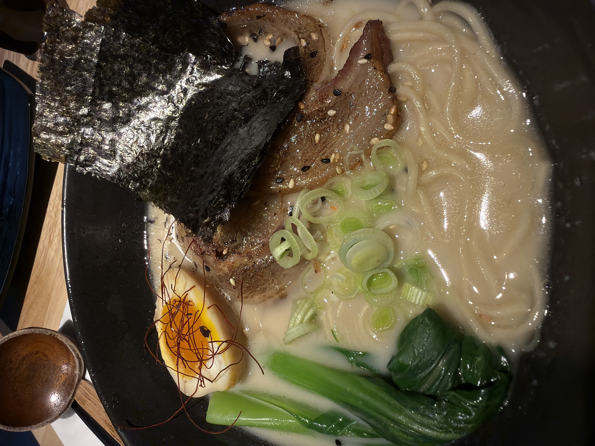 A top-down, eye-level shot captures a bowl of ramen. Prominently featured are two pieces of seaweed and sliced pork with sesame seeds. Chopped green onions, bok choy, and a half-cut soft-boiled egg decorated with chili threads are also visible. The broth appears creamy, and the noodles fill the remainder of the bowl. The bowl itself is dark, and a small wooden spoon sits beside it. The scene is set on a wooden table. There is no indication of the location being North Kuta, Indonesia. No people are visible.