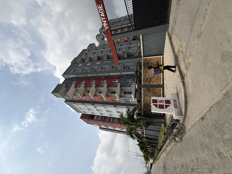 The scene depicts an urban environment in Igboefon, Nigeria, under a partially cloudy sky. A multi-story residential building, approximately seven to eight floors tall, dominates the right side of the image. Its exterior features a modern design with alternating sections of grey and red facades, numerous white-railed balconies, and visible air conditioning units. The property is enclosed by a dark grey perimeter wall topped with barbed wire. An overhead red and grey archway at the entrance bears the word "VINTAGE" in red capital letters. Adjacent to the entrance, a white concrete monument sign with a red geometric logo displays the word "STELLAR."
In the foreground, a paved street with grey interlocking bricks extends to the left, bordered by a concrete drainage channel. A single individual with dark skin walks on the paved sidewalk towards the right. They wear a yellow long-sleeved shirt over a dark garment, dark trousers, and dark shoes, carrying a purple delivery bag on their back and holding a dark object, likely a mobile phone, in their right hand. A black bicycle is parked upright against the curb on the left, near the "STELLAR" sign. Several palm trees are visible lining the property fence. Streetlight poles are discernible further down the road on the left.