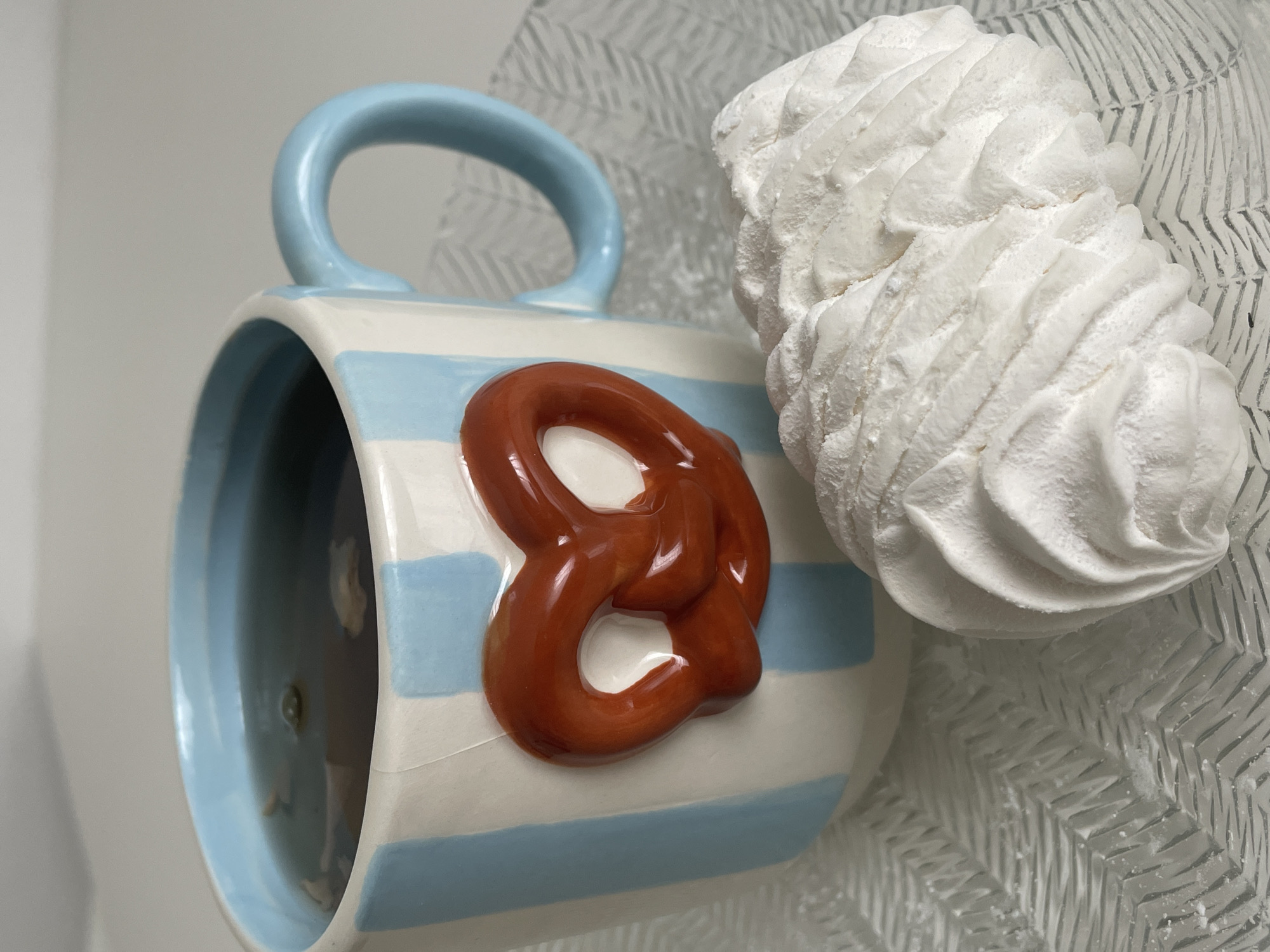 The image shows a close-up view of a white mug with blue stripes, decorated with a three-dimensional red pretzel on the front. The mug is filled with a dark liquid, possibly tea, with some light-colored pieces floating in it. Next to the mug is a large, white, fluffy pastry, possibly a marshmallow or zephyr. Both the mug and the pastry are placed on a textured, clear glass plate with a zigzag pattern. The background is a plain white surface. There is no indication of the city of Nidda, Germany, or any people in the image.