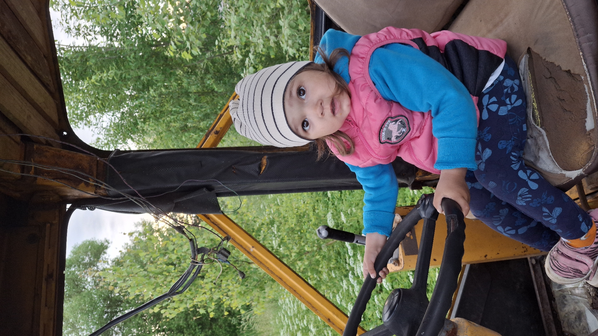 A young child, likely a girl, is the sole subject in this media file. She is seated in the driver's seat of what appears to be an old, yellow tractor. The child is wearing a striped beanie, a bright blue long-sleeved shirt, and a pink quilted vest. She also has on dark blue leggings adorned with a blue butterfly pattern and pink sneakers. Her right hand is gripping the steering wheel, suggesting she is engaged in playing with the tractor.
The setting is outdoors, with lush green trees and foliage visible in the background. The lighting suggests it is daytime, possibly late afternoon given the soft quality of the light. The tractor appears weathered and possibly abandoned, with exposed wires and worn upholstery on the seat.
The child's expression is one of innocent curiosity and engagement, with her eyes wide and looking directly at the camera. The overall mood is one of childhood play and exploration in a rustic environment. There is a visible patch on the child's vest with text that appears to say "TEAM HORSE".