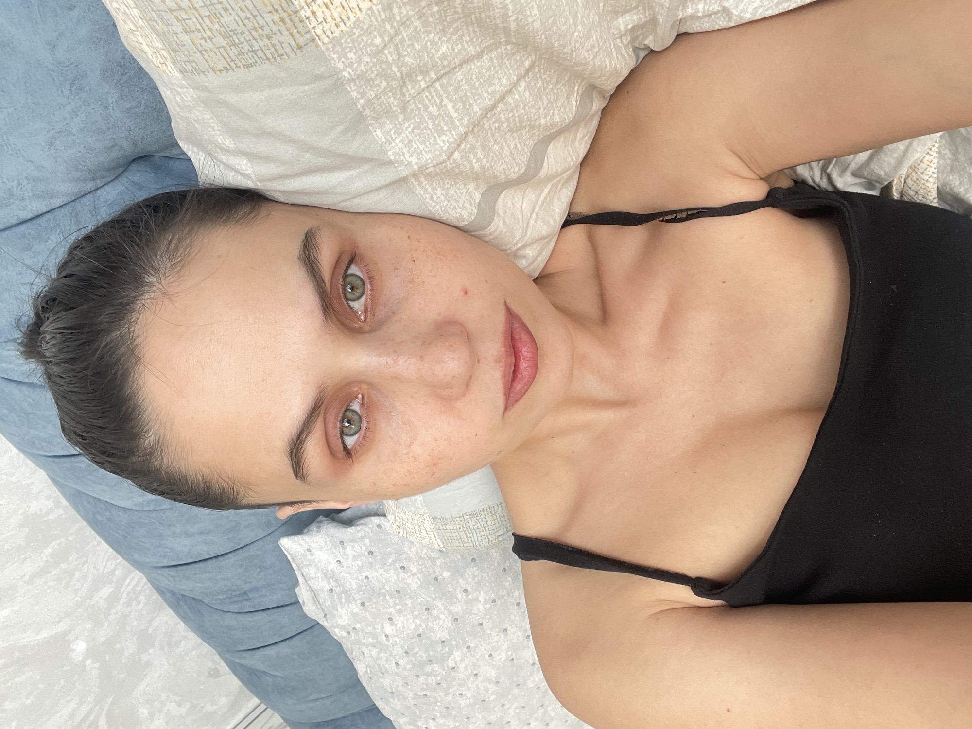 The image shows a young woman lying down indoors, likely on a bed or a comfortable sofa, in Izhevsk, Russia. She is taking a high-angle selfie, looking directly at the camera with a calm and relaxed expression. Her dark hair is pulled back, and her face is natural, featuring prominent freckles across her nose and cheeks. She has striking green eyes and her lips are slightly parted. She is wearing a simple black spaghetti strap top. Her head rests on a light-colored, patterned pillow, with a blue-grey upholstered surface visible to her left. The lighting is soft and even, suggesting a daytime setting indoors. No text is visible in the image.