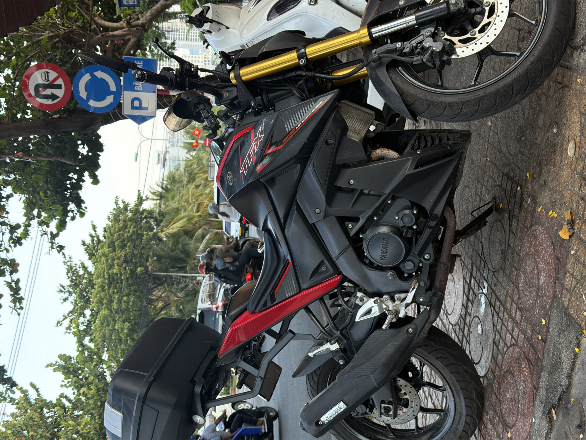 The image shows two parked motorcycles on a paved street in what appears to be a populated area. The motorcycle in the foreground is a black and red Yamaha with a black storage box attached to the rear. The other is white. Traffic signs indicating "No U-turn" and a traffic roundabout direction are visible in the upper left corner. Trees are present, obscuring the view of other vehicles and buildings in the background. The background suggests that the location is in Nha Trang Ward, Vietnam.