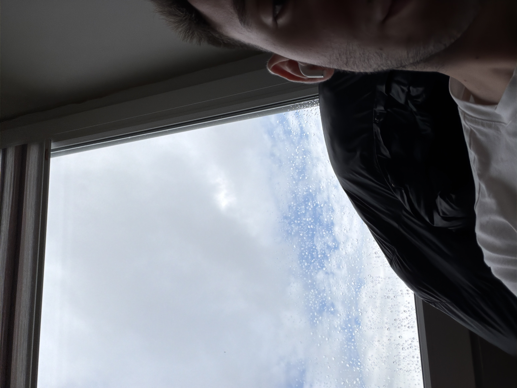 The image, captured from a low-angle perspective, features a person partially visible in the upper right and a window providing a view of the sky. The person's head and upper torso are visible, including brown hair, an eye, an ear with a white earbud, a jawline with stubble, and part of the neck. They are wearing a white t-shirt underneath a black, possibly waterproof, jacket.
A prominent light grey window frame horizontally bisects the image. To the left, vertical striped brown curtains or blinds are partially visible. The window pane is distinctly covered in numerous raindrops, indicating recent or ongoing rain. Beyond the window, the sky is overcast with a mix of light grey and white clouds, with some patches of blue sky showing through.
The scene depicts a rainy or recently rainy daytime, with the person seemingly looking out of the window or capturing the weather conditions in Sofia, Bulgaria. There is no visible text in the image.