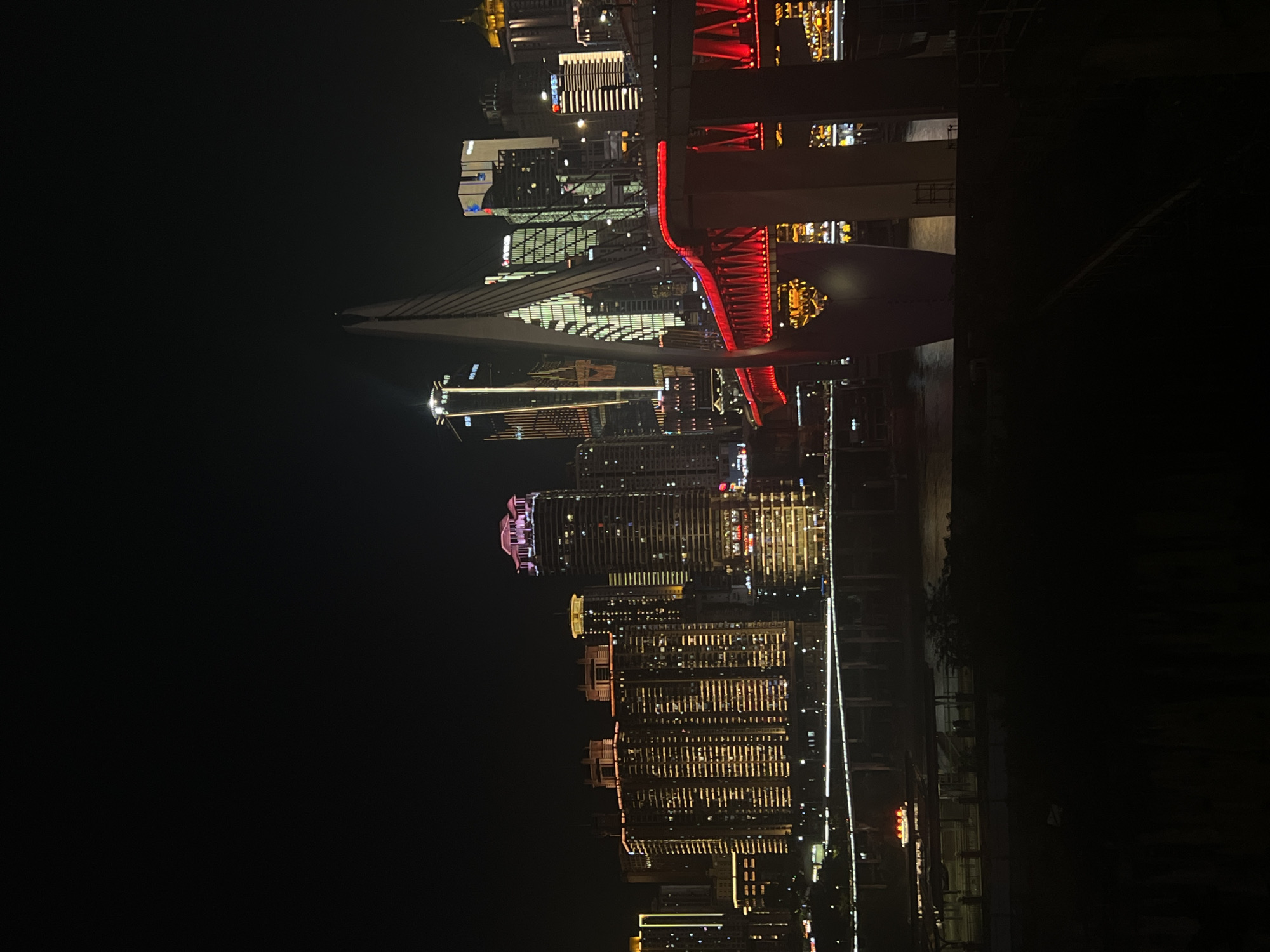 The image shows a nighttime view of a cityscape, likely in Jiangbeicheng Street, China, dominated by illuminated skyscrapers and a brightly lit bridge. The foreground is dark, with silhouettes of vegetation or structures. A multi-tiered bridge, illuminated with vibrant red lights along its structure, spans across a body of water. Behind the bridge, a cluster of tall buildings rises, their windows glowing with various colors, predominantly warm yellow and white. Some buildings have distinctive architectural lighting, such as the pink-capped building on the far right and the angular, illuminated facade of a skyscraper to its left. A distinctive cable-stayed bridge tower, white and pointed, is visible in the mid-ground. The water reflects the city lights. In the lower left portion of the frame, a small boat with yellow lights is visible on the water, near a dock area. The overall scene depicts a lively and illuminated urban environment at night.