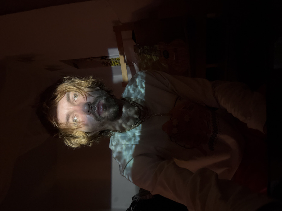 A low-lit indoor shot features a person with light brown hair and a beard. The person wears a light-colored, long-sleeved shirt and a necklace. A pattern of light illuminates part of their face and clothing. In the background, blurry objects suggest a shelf or room with various items. The lighting creates a strong contrast between light and shadow.