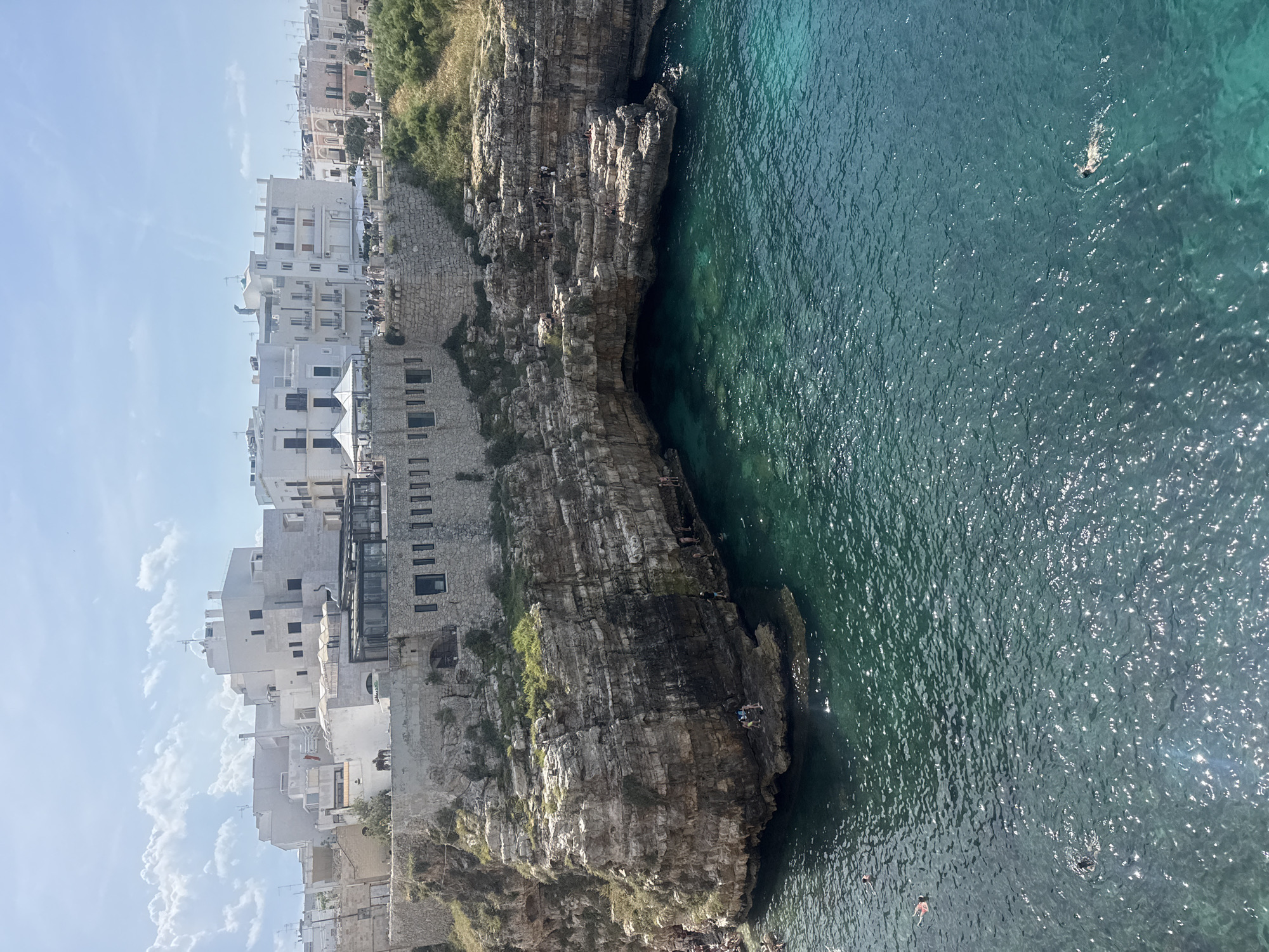Here is a detailed description of the image:
The image shows a scene in Polignano a Mare, Italy, featuring a rocky coastline and turquoise sea. The camera angle is elevated, offering a view down towards the water. The water is textured with gentle ripples, indicating calm conditions, with several people swimming and sunbathing.
Adjacent to the water is a rugged cliff face, with exposed rock layers and patches of vegetation. Some people are sitting on the cliff.
On the left side of the image are closely built white buildings, a characteristic feature of Polignano a Mare. Some of the buildings appear to be built directly onto the cliffs, merging the man-made structures with the natural landscape. The sky is light blue with a few white clouds.