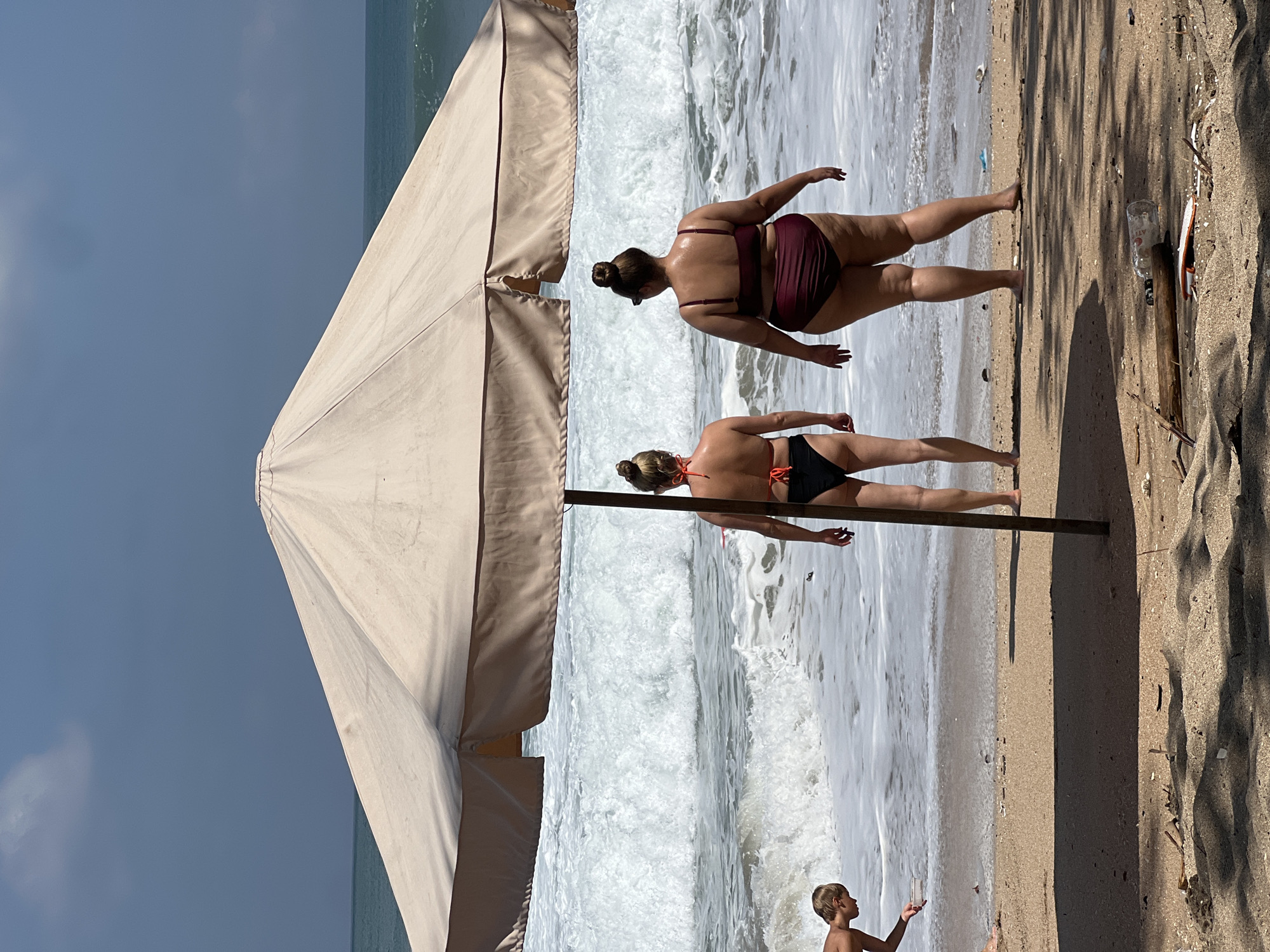 The image depicts a beach scene in Kuta, Indonesia. Two women are shown in bikinis, standing near the ocean. The woman in the foreground is wearing a black bikini, while the woman behind her is in a maroon bikini. Both have their backs to the camera. In the lower right corner, a young boy is visible, looking out towards the water. A large beach umbrella partially covers the scene on the left. The ocean is active with waves breaking on the shore. The sand appears light brown, and shadows are cast from the umbrella and possibly palm trees, suggesting it is daytime. The sky is a clear blue. A plastic bottle and some debris are visible on the sand. The overall mood is relaxed and summery, typical of a beach environment.