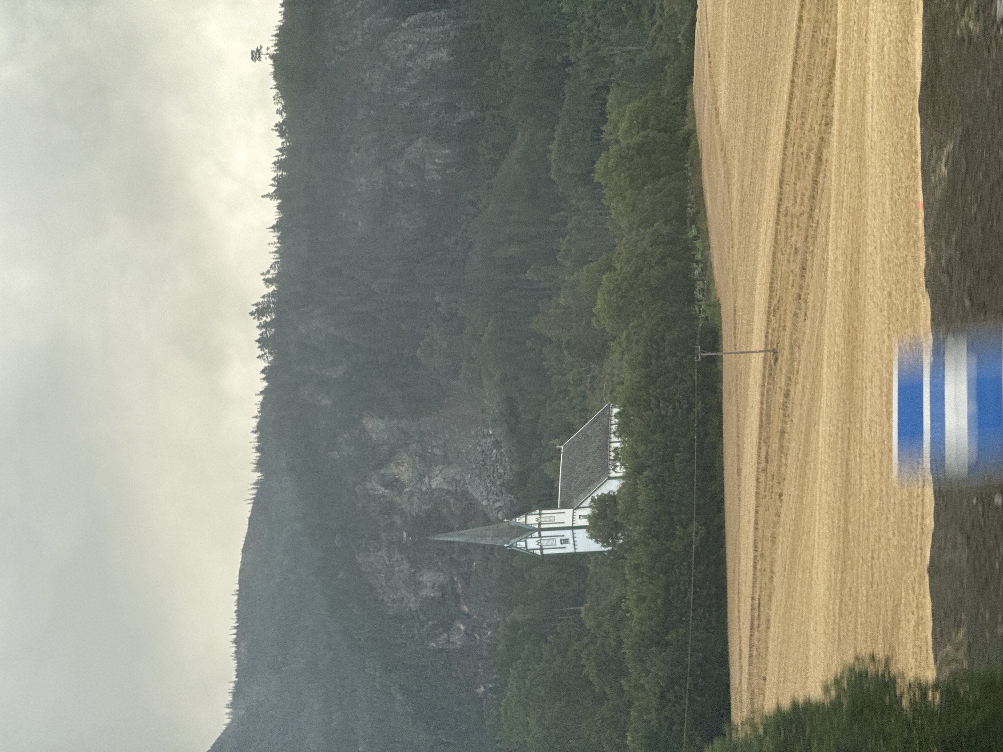 The image shows a white church with a tall spire nestled amongst dense green trees, situated on a hillside. Behind the trees, a rugged, rocky mountain face rises, and the summit is dotted with evergreen trees. A large field of harvested, golden-brown crops lies in the foreground. The sky is overcast with a grey, cloudy atmosphere. In the bottom right corner, a blurred blue and white object suggests motion, possibly from a vehicle. This scene is characteristic of the rural landscape found in the vicinity of Skatval, Norway, a coastal village known for its natural beauty and historical sites.