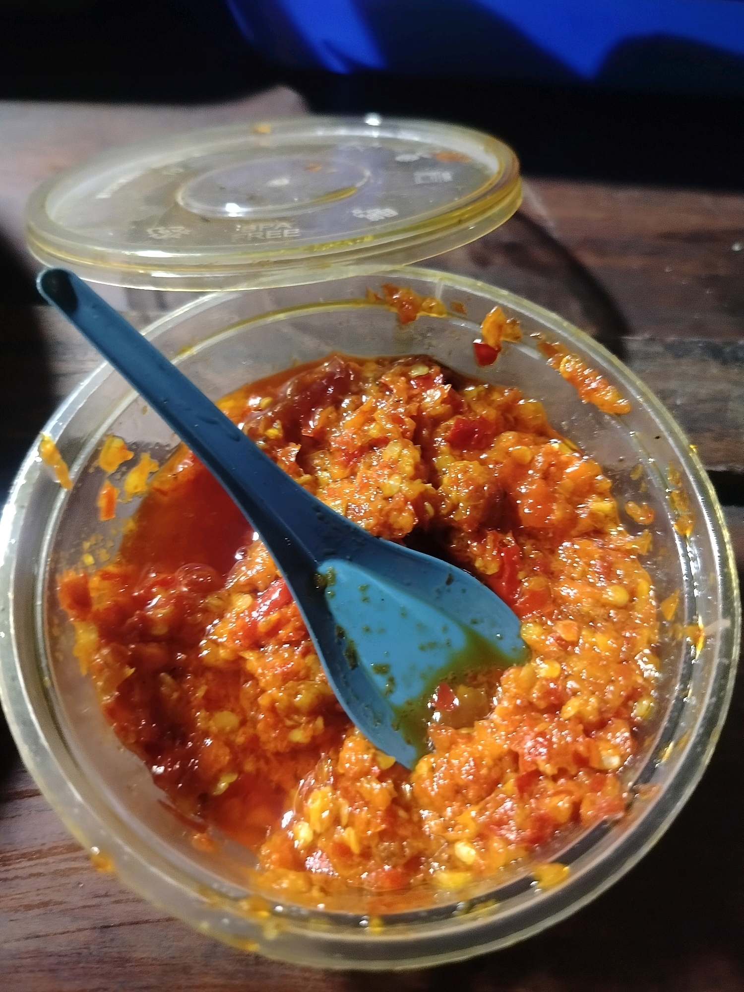 A close-up, overhead shot shows a bright red, chunky chili paste in a clear plastic container. A light blue plastic spoon is partially submerged in the paste, with its handle angled out of the container. The container has a clear plastic lid, partially ajar, resting behind it. The lid has some markings on it, including "BPA FREE". The container and lid are sitting on a wooden surface. The background is dark, with a blurred blue object visible in the upper right. The image has a slightly grainy quality. The location is not explicitly visible in the image.