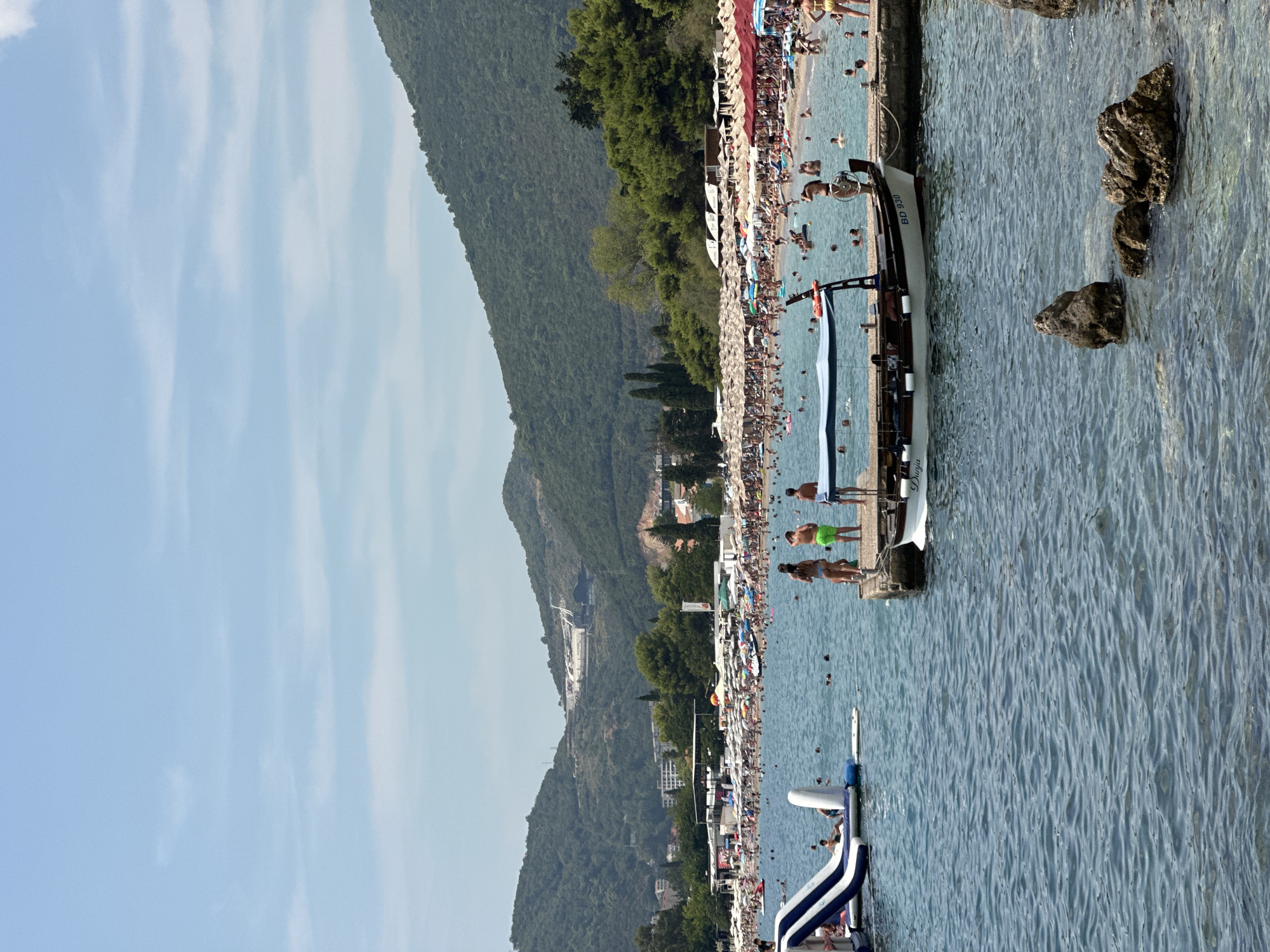 The image depicts a crowded beach scene in Budva, Montenegro, on a sunny day. The sea is calm with visible ripples. In the foreground, a small boat with the registration "BD 930" is docked at a pier. Several people are standing on the pier, including a woman in a bikini and two young boys. To the left of the pier, an inflatable water slide is visible in the water, with a few people around it.
The middle ground is dominated by the beach, packed with numerous sunbathers relaxing under a multitude of umbrellas. The water is dotted with swimmers. In the background, lush green hills rise, characteristic of the Montenegrin landscape, with some buildings and structures visible on their slopes. The sky is a clear, pale blue with a few wispy clouds. The overall impression is a lively summer day at a popular tourist destination.