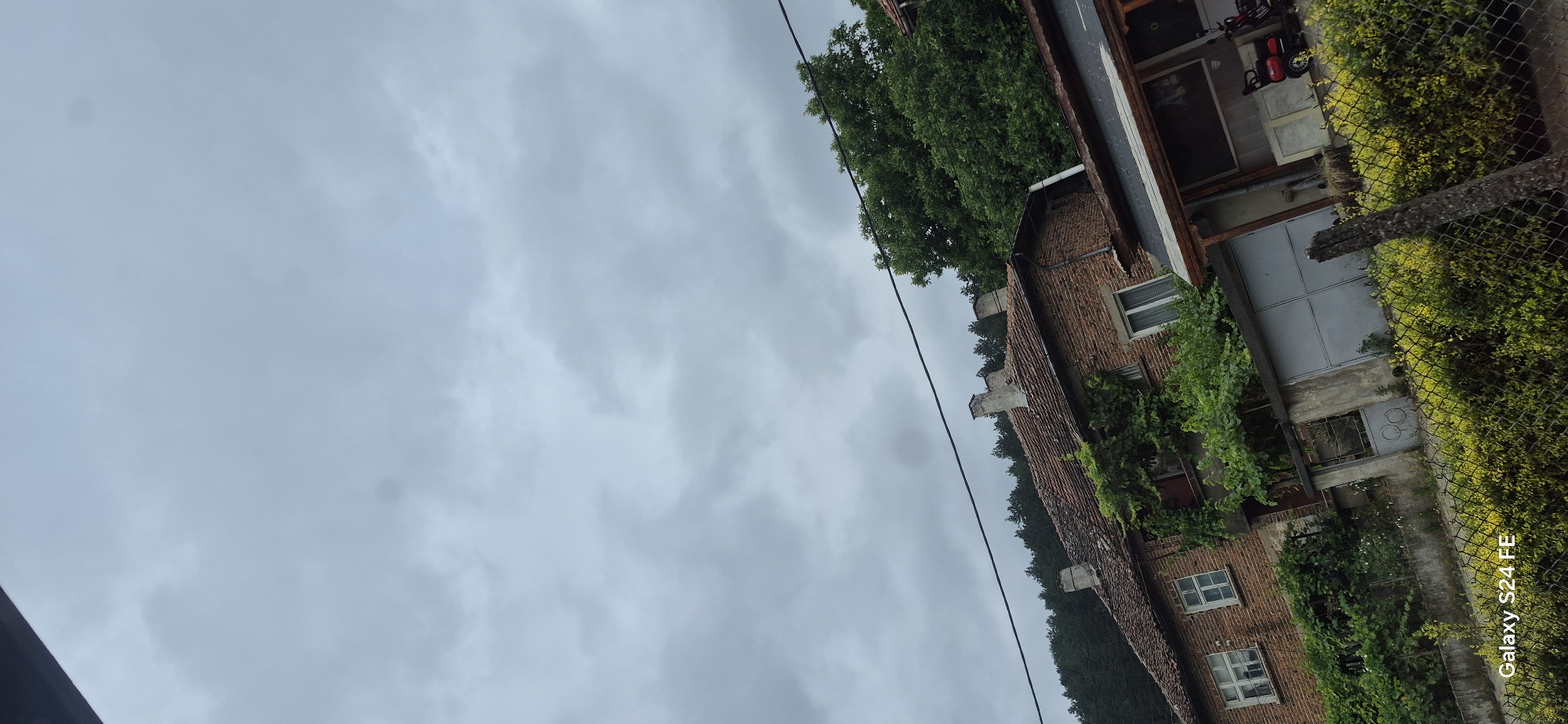 The image depicts a rural scene in Malko Tarnovo, Bulgaria, under a cloudy sky. In the foreground, a wire fence with dense green foliage is visible. Behind the fence, a weathered brick house with a tiled roof and several chimneys stands out. Green vines and plants climb the side of the house, adding to its rustic appearance. A grey metal gate is partially visible, leading to a courtyard. Adjacent to the house is a covered outdoor area with a wooden structure, where a red motorized scooter is parked. Further in the background, a dense forest of pine trees covers a hillside, characteristic of the Strandzha Mountains region where Malko Tarnovo is located. The overall atmosphere suggests a quiet, possibly damp day in a village setting.