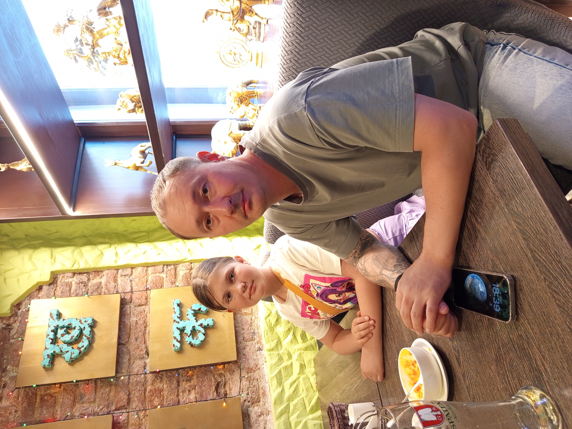 A man and a young girl are seated at a wooden table in what appears to be a restaurant. The man, on the right, is wearing a grey t-shirt and blue jeans. He has a tattoo on his left arm. The girl, on the left, is wearing a white t-shirt with a colorful graphic and a yellow crossbody bag. She has blonde hair pulled back in a ponytail. In front of them are a glass of beer, a bowl of food, and a smartphone. The background shows a brick wall with decorative turquoise symbols and shelves filled with golden figurines. The lighting suggests an indoor setting.