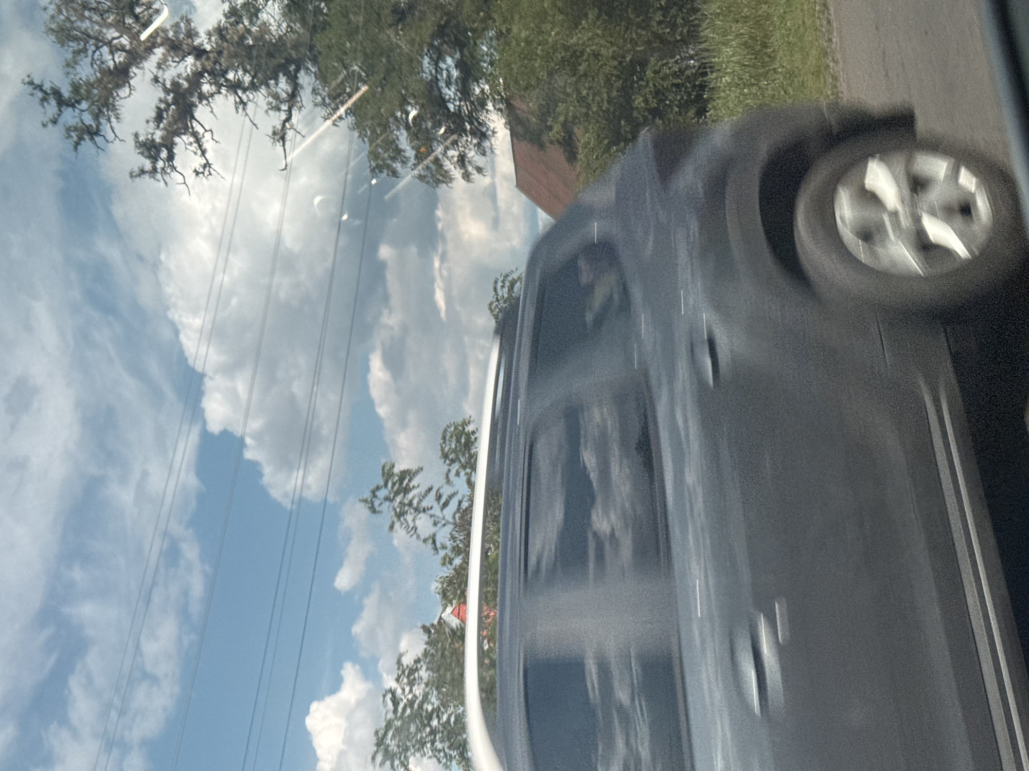 The image depicts a scene observed from a moving vehicle, likely in Markivka, Ukraine, during daylight hours. A dark grey mid-size SUV, equipped with white roof rails, is the primary subject, shown in motion from its passenger side and rear quarter, traveling from left to right. Significant motion blur is evident on the vehicle's body and its multi-spoke alloy wheels, which appear as blurred circular shapes. Reflections of the sky and clouds are visible on the SUV's darkened windows.
The background consists of a bright blue sky with scattered white clouds, including cumulus formations and wispy cirrus clouds. Multiple dark utility lines, appearing as five parallel horizontal strands, traverse the sky in the upper and middle portions of the frame. Various trees are visible along the roadside; on the left, blurred green foliage is present, while on the right, a large tree with pronounced, gnarled bare branches stands against the sky. Further right and partially obscured by trees, a building with a distinct red, gabled roof is discernible. The immediate foreground on the right side of the road shows dark asphalt and green roadside vegetation. Several bright, streak-like lens flares and circular reflections are present, particularly in the upper right quadrant of the image, consistent with light interacting with a car window or camera lens. No specific individuals are clearly identifiable within the frame.