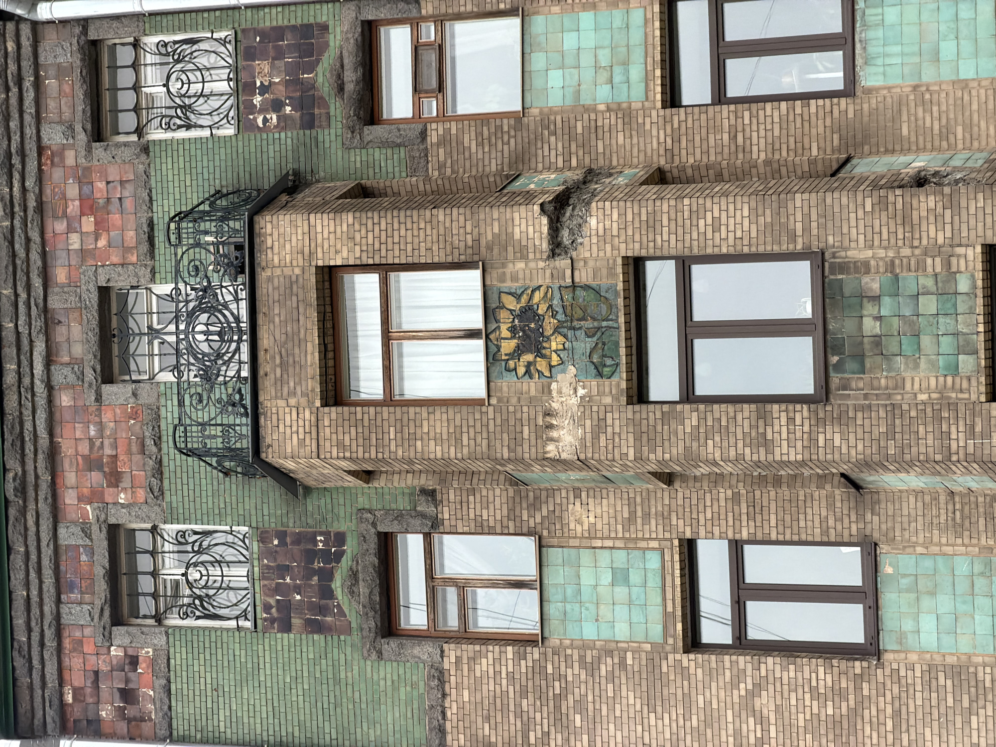 The image depicts the facade of a building in Saint Petersburg, Russia, notable for its unique architectural style. The building is constructed with a variety of materials, including brick, tile, and stone. The facade is adorned with windows of varying designs, some with decorative ironwork. A small balcony is visible, also featuring elaborate iron detailing. The exterior is further embellished with tiled panels in different colors, including green, brown, and red. A prominent decorative sunflower design is set between windows. The building shows signs of age and wear, including areas of damage and repair. The overall composition suggests a blend of architectural styles, typical of buildings constructed during the Art Nouveau period.