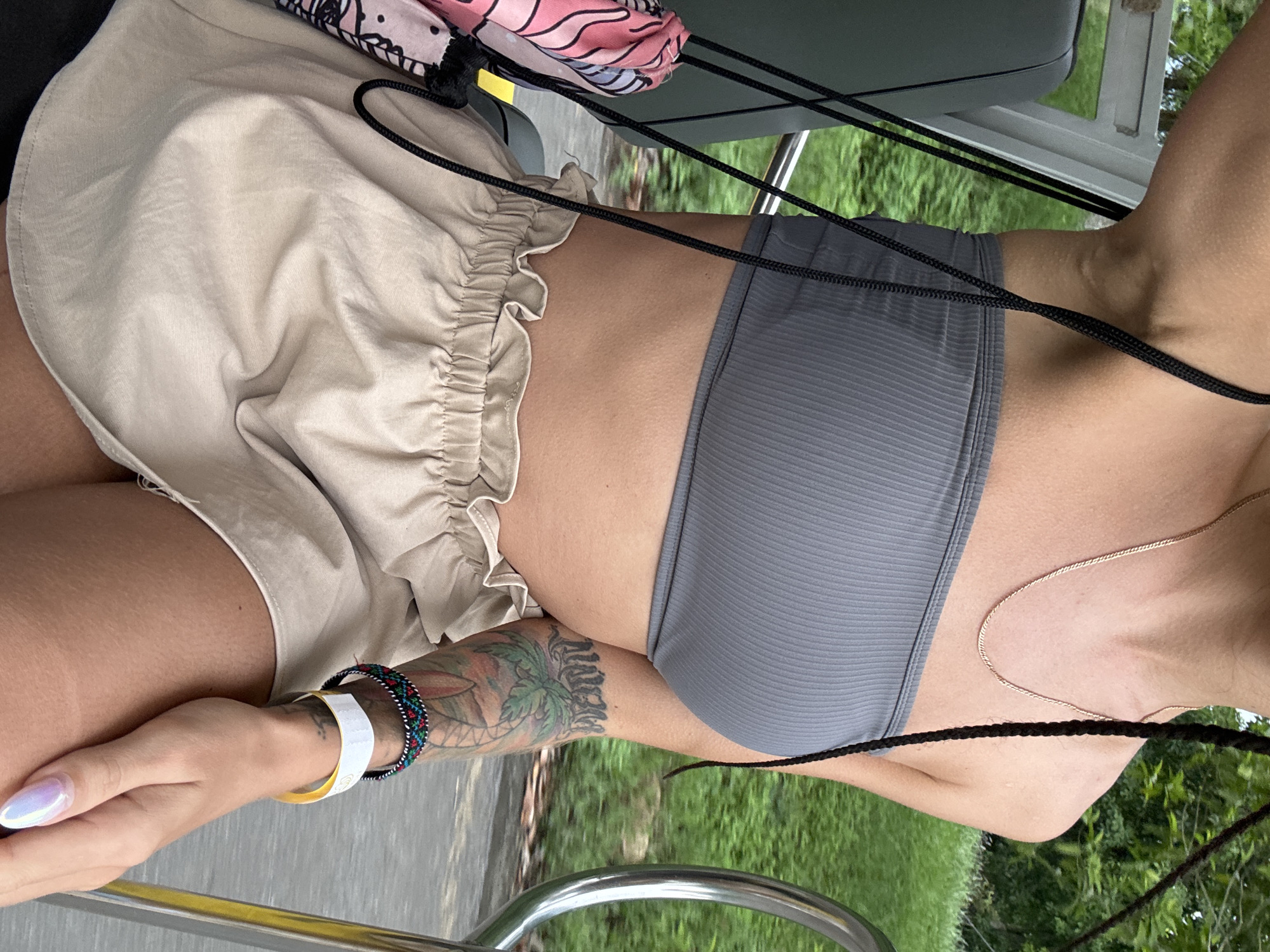 The image shows a close-up of a person's torso and arms, focusing on their clothing and accessories. The person is wearing a grey ribbed bandeau top and a tan ruffled skirt. A portion of a colorful scarf with a pink and black pattern is visible on the left side of the frame, attached by black cords. The person has a tattoo on their right arm, which appears to depict a palm tree and lettering. On their wrist, they are wearing a patterned bracelet and a white wristband. The background consists of blurred green foliage, suggesting an outdoor setting. The image is shot from a slightly elevated angle, looking down at the person.